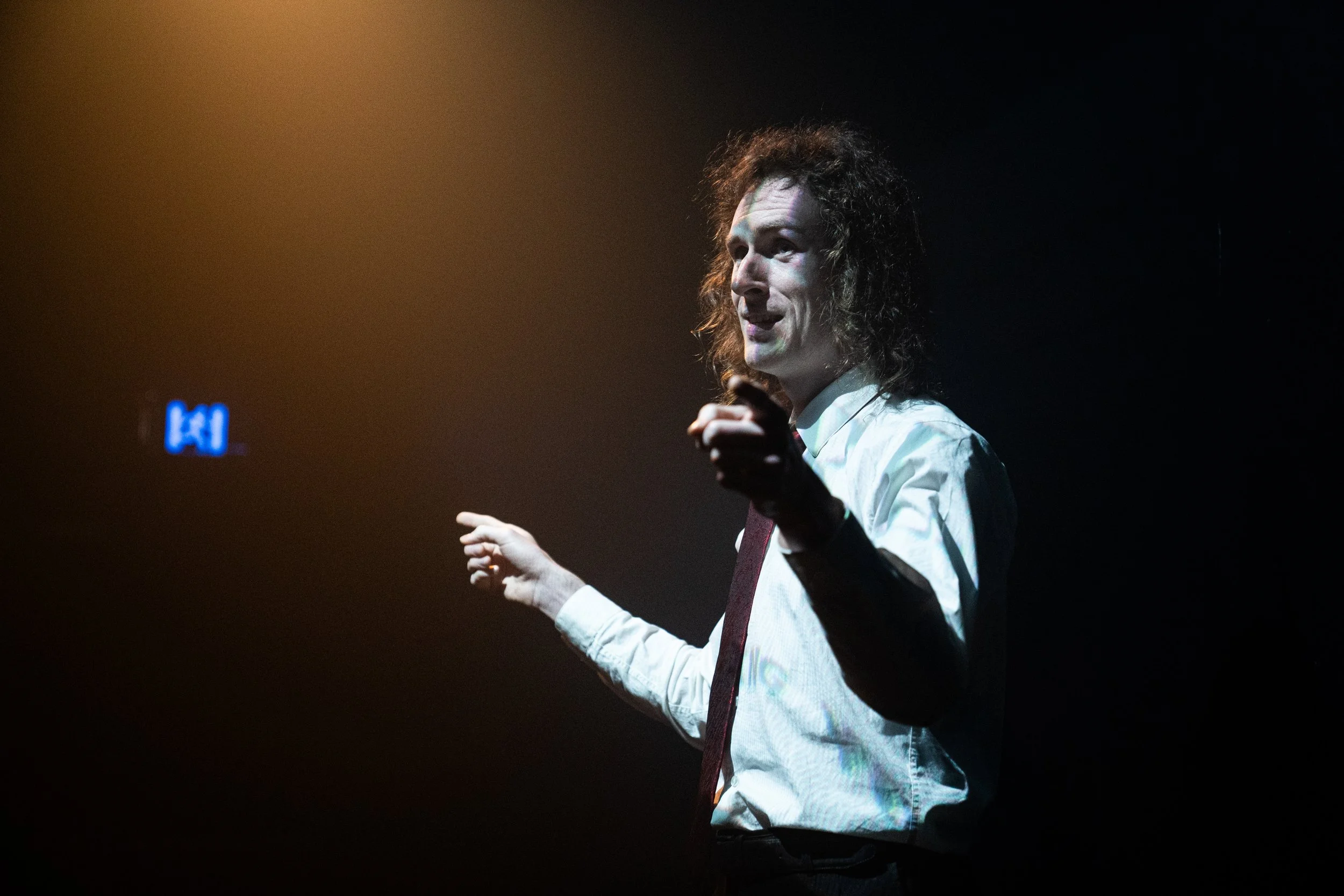Person with curly hair in a white shirt and tie, speaking or presenting on stage with a dark background and some stage lighting.