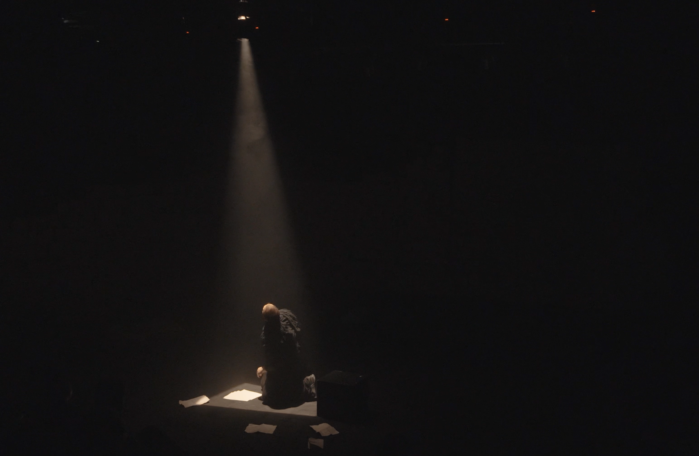 A person kneeling on the ground in a dark space, illuminated by a spotlight from above, surrounded by scattered papers.