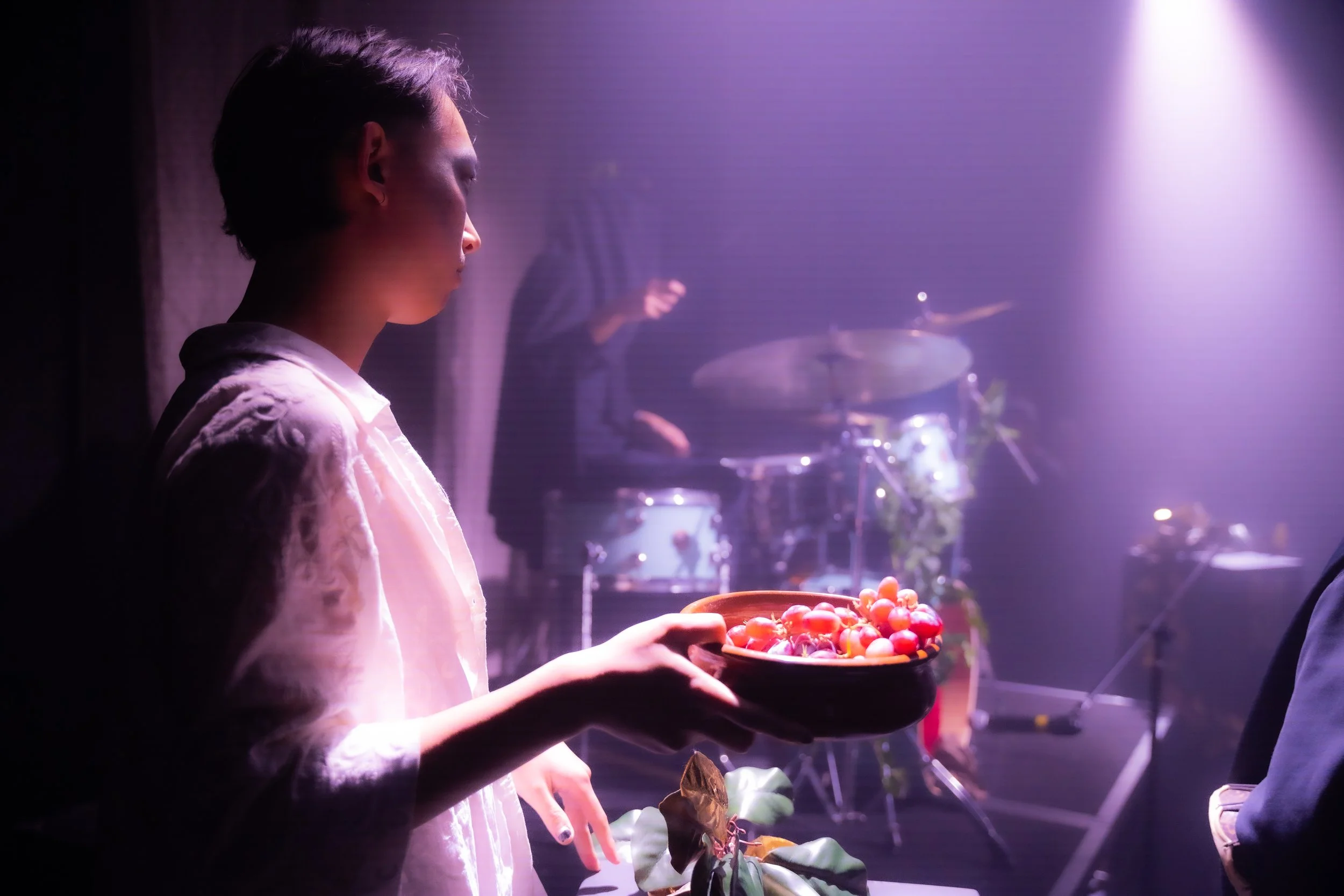 A young woman holding a bowl of cherry tomatoes on stage with band equipment and a drummer in the background, illuminated by purple stage lights.