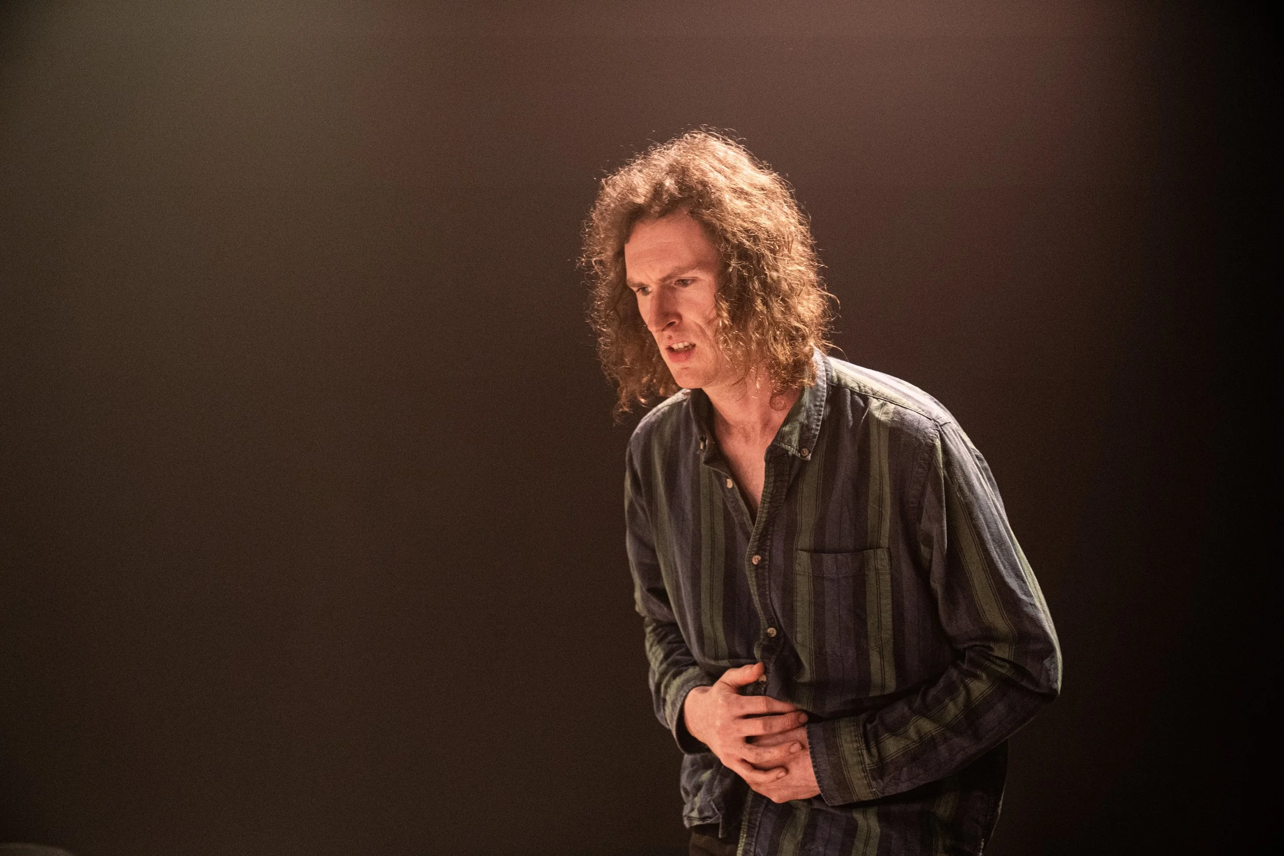 A man with curly hair and a striped shirt, appearing to be in pain or discomfort, holding his abdomen.