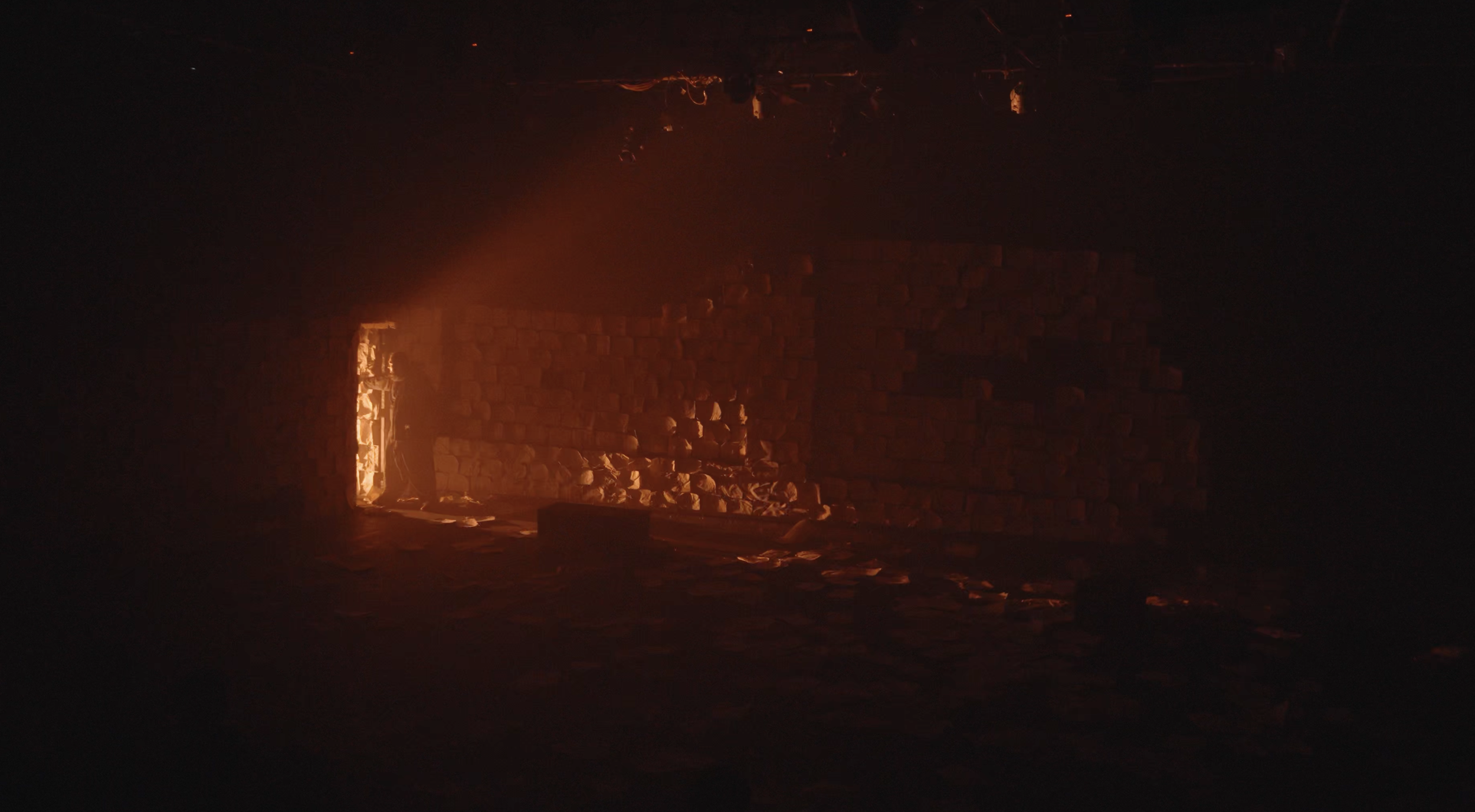 A dark room with an open door emitting a warm orange glow. Inside, stacked bricks can be seen with some debris on the floor.