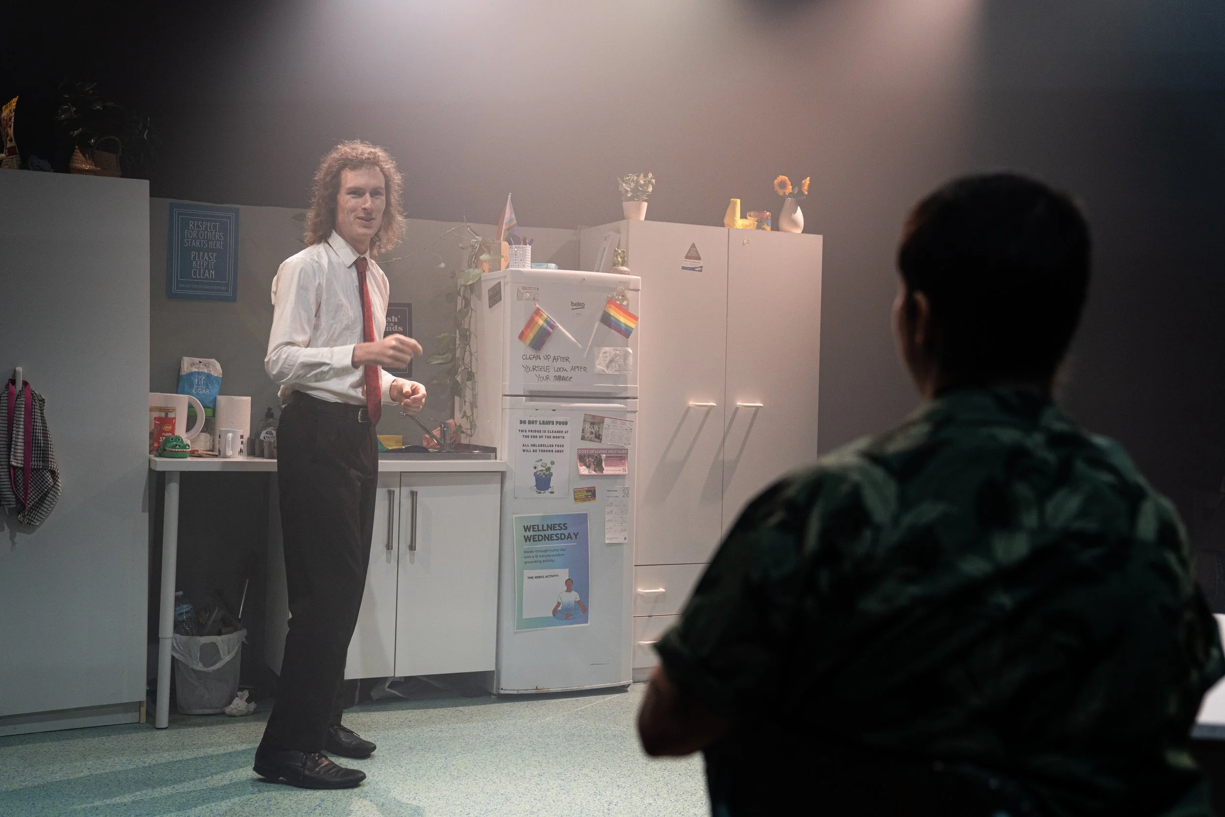 A person with curly hair wearing a white shirt and red tie standing in a kitchen, talking to another person in camouflage clothing. The kitchen has a white refrigerator decorated with rainbow flags, a white cabinet, and various items on top like vase