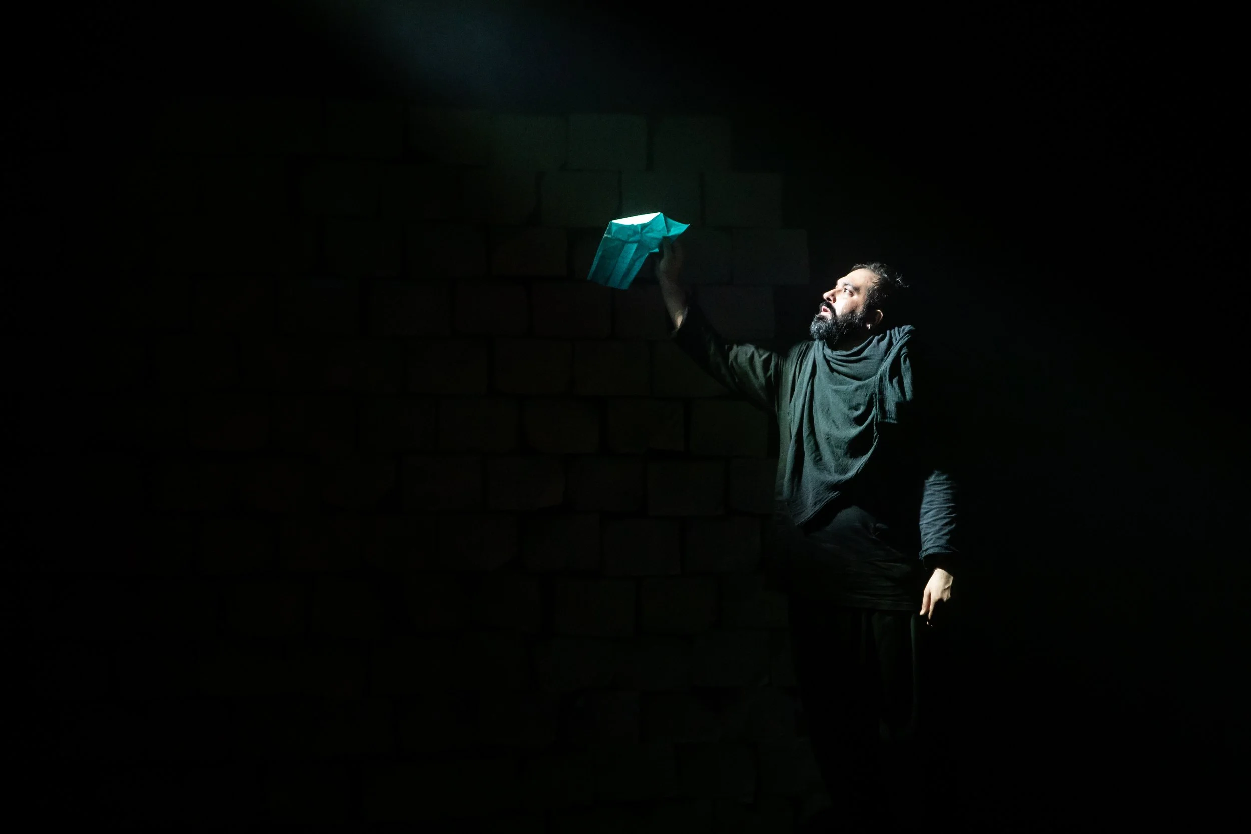 Man with a beard, dressed in black, holding a blue paper bag, illuminated by a spotlight in a dark space with a brick wall background.