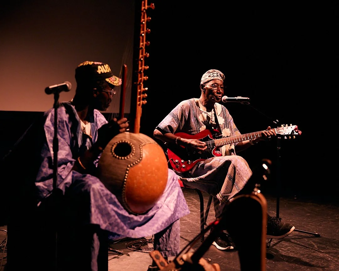 Two musicians performing on stage, one playing an acoustic guitar and singing into a microphone, the other playing a traditional African drum.