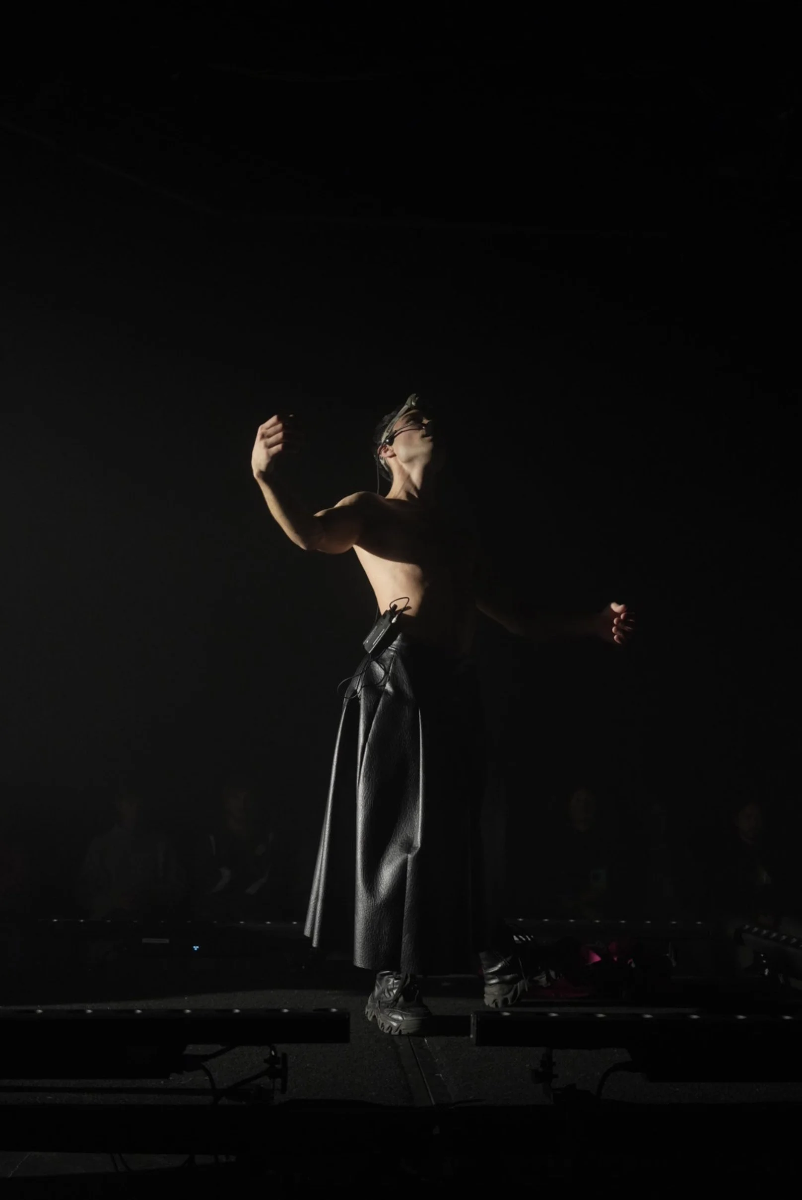 A shirtless performer on stage wears black pants, sneakers, and a headband, with hands raised and eyes closed, illuminated by a spotlight in a dark setting.