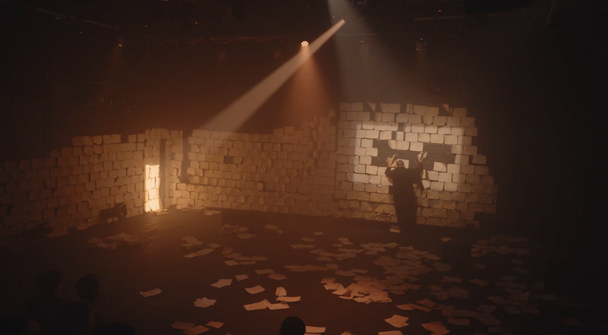 A stage set with a wall made of stacked bricks and scattered papers on the floor. A person is pressing against the wall through a hole, with a spotlight shining down from above.