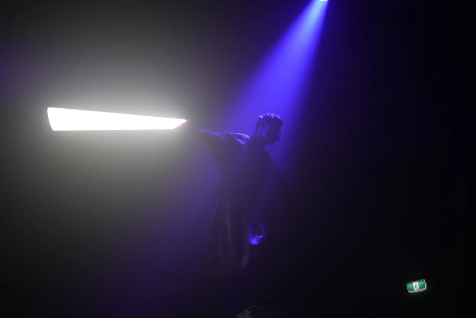 A person silhouette illuminated by a bright white light and blue stage spotlight, standing with arms outstretched in a dark setting.