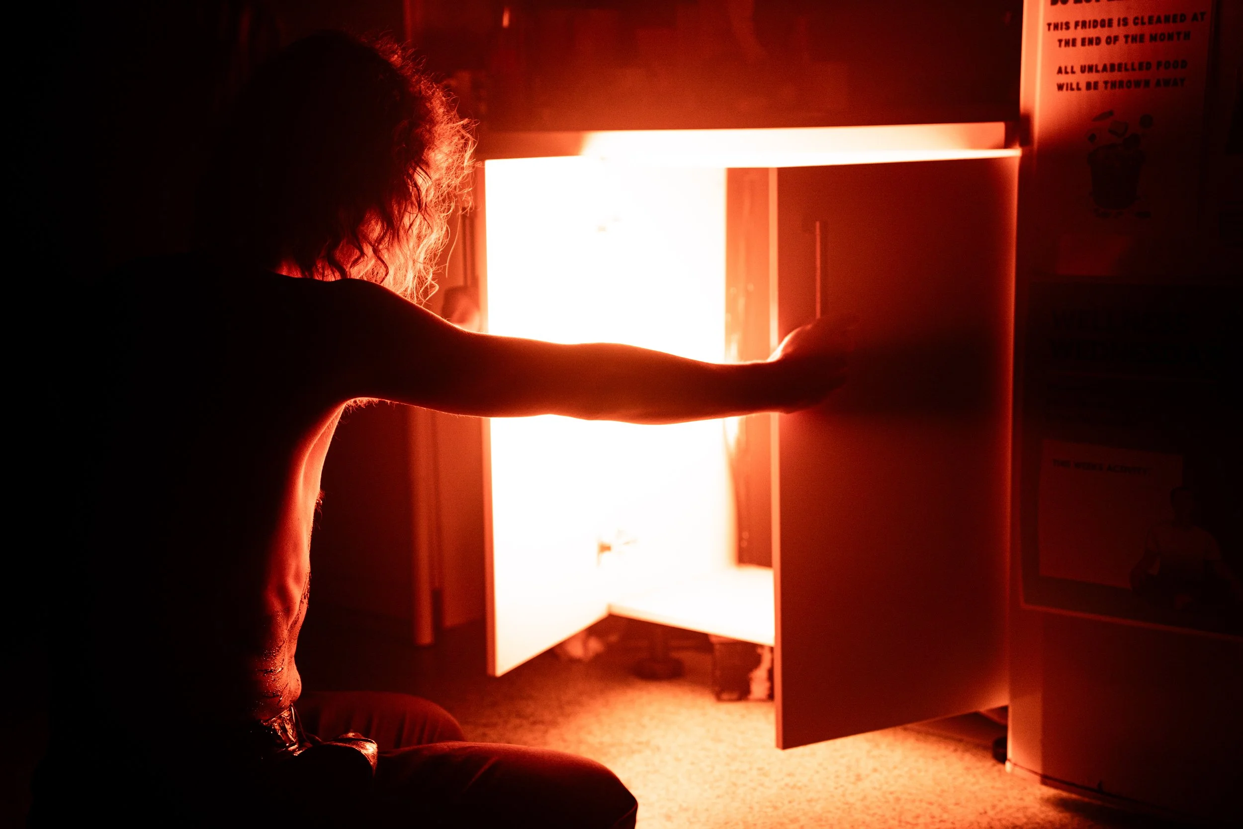 A person with curly hair reaching towards an open fridge with bright light inside, in a dark room.