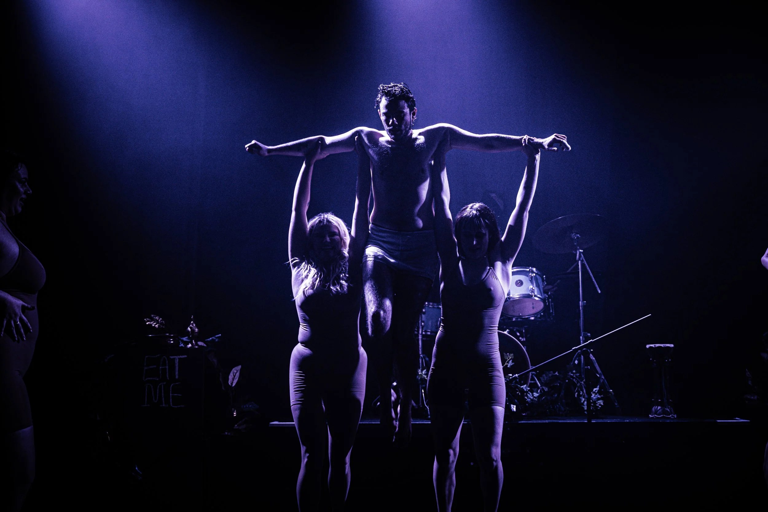 A man is being lifted in a crucifix pose by two women on a dark stage with blue and purple lighting, with musical instruments in the background.