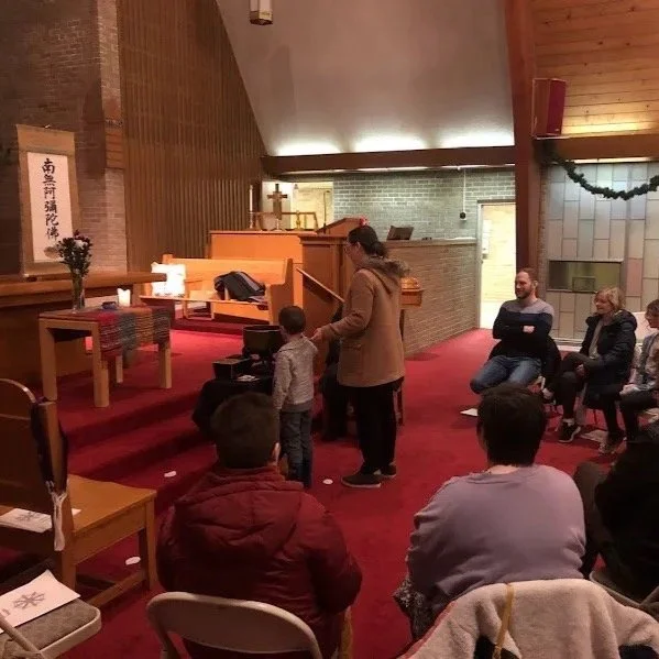 A woman and a young boy are standing in front of a small congregation inside a church. The woman appears to be speaking or reading while the boy stands beside her. The congregation is seated, listening attentively. The church interior features wooden walls, a red carpeted floor, and an altar in the background with candles and a cross. Some people are smiling and watching.</imageAltText>