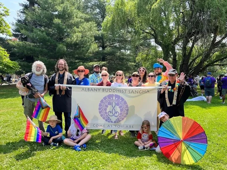 Group of people holding a banner that reads 'Albany Buddhist Sangha' and 'Come As You Are,' outdoors on a sunny day with trees, rainbow flags, and a rainbow umbrella, celebrating diversity and inclusion.