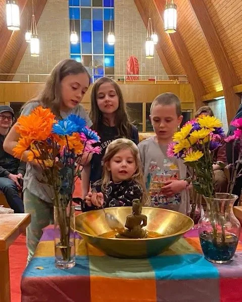Children gather around a table with a golden bowl containing a small Buddha statue, surrounded by colorful flowers in vases inside a church.
