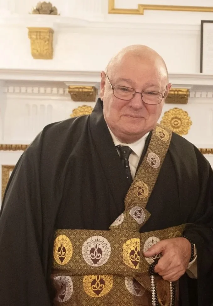 A man in formal religious attire holding a beaded necklace, standing in front of a decorative background with gold accents.