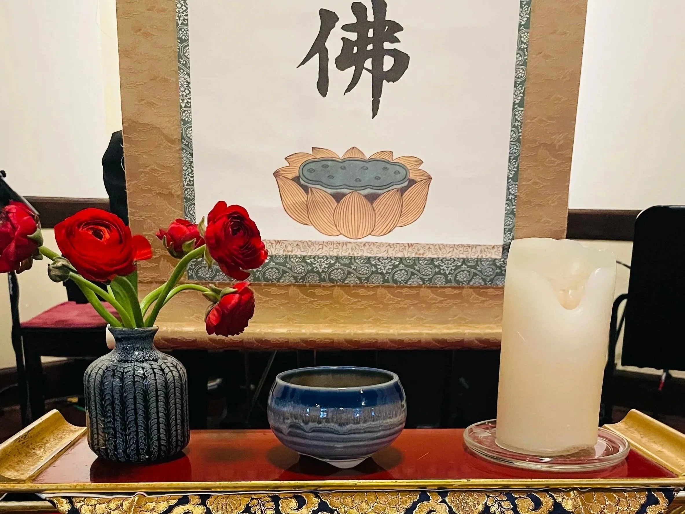 A table with a black and white ceramic vase holding red flowers, a blue and white ceramic bowl, and a large white candle on a glass holder. In the background, a decorative scroll with Chinese characters and an illustration of a lotus flower surround the calligraphy.