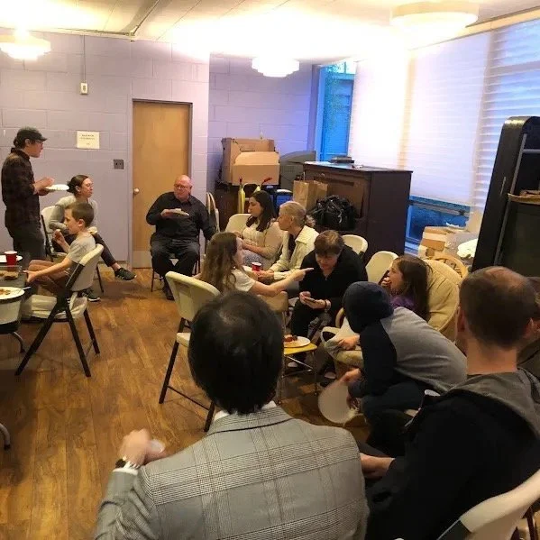 Group of people seated in a room having a discussion or meeting, with a person standing and speaking to the group, some holding cups and papers, in an indoor setting.