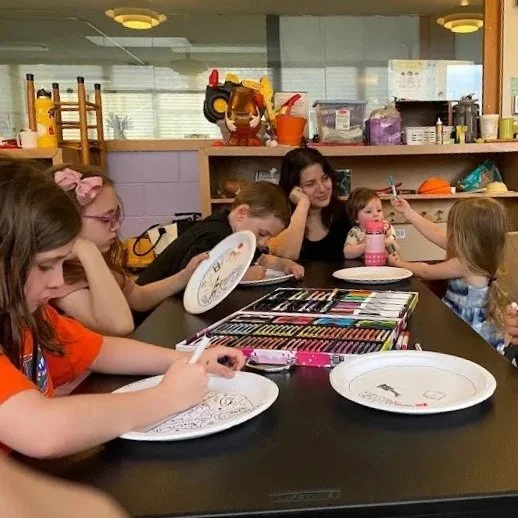 Children and a woman sitting at a table coloring on paper plates with markers, in a room with shelves and toys in the background.
