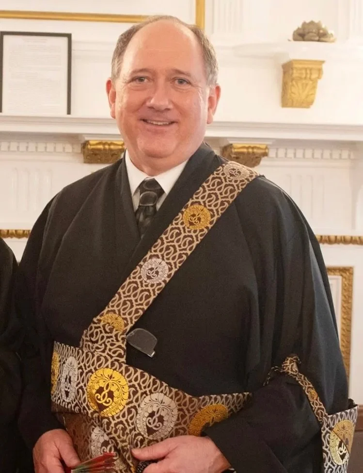 A man dressed in a graduation gown and stole, smiling, standing in a room with white and gold decor.