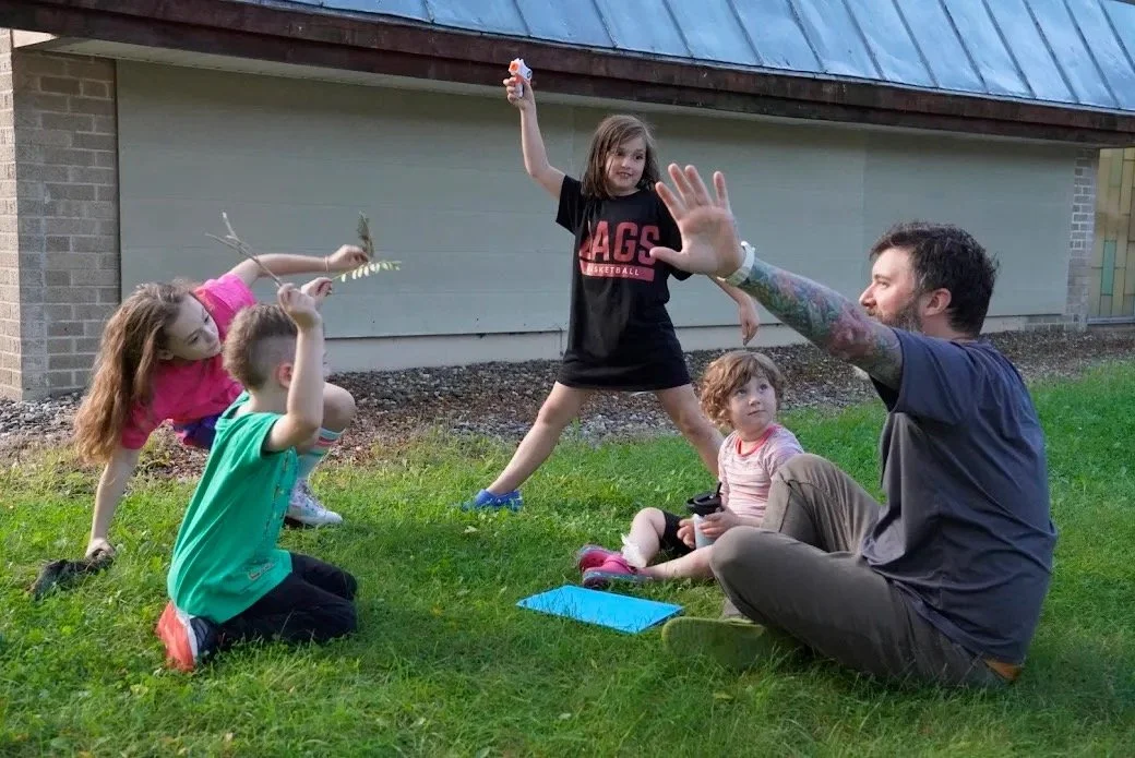 Children playing with a man outdoors on grass, one girl holding a branch, others sitting and standing, in front of a building with a brick wall and roof.