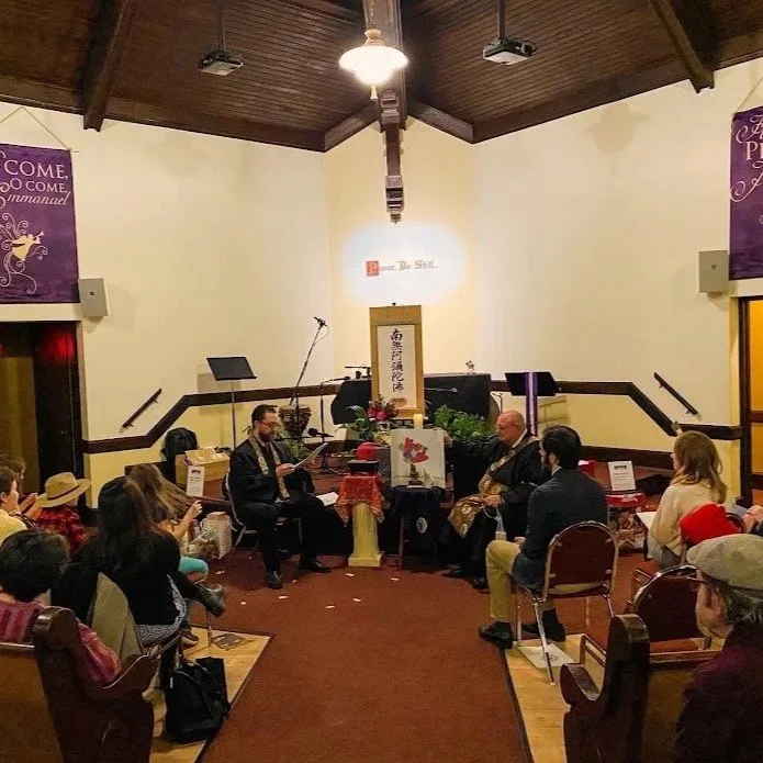 A small cultural or spiritual gathering in a room with a wooden ceiling and beige walls. People are seated in a semi-circle, listening to two men seated in front of a table with decorations and a framed calligraphy scroll. The setting appears to be a lecture or presentation, with a piano and flowers on the table.