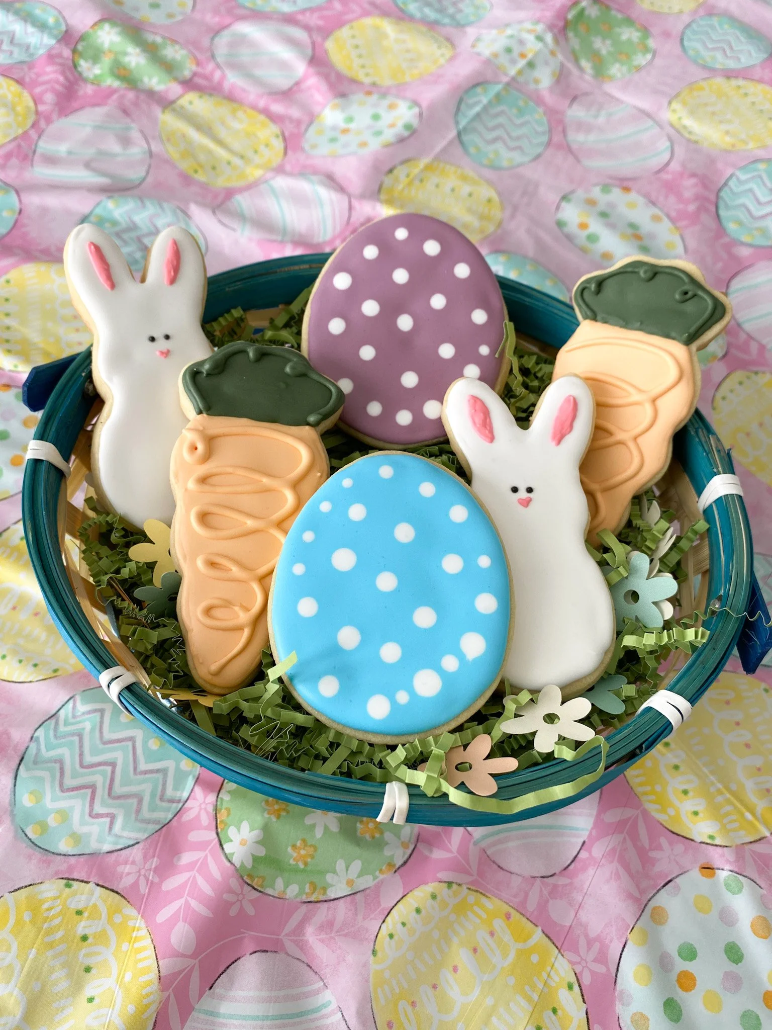 Easter-themed sugar cookies in a basket, including bunny, carrot, and decorated egg shapes, with a colorful egg-patterned cloth underneath.