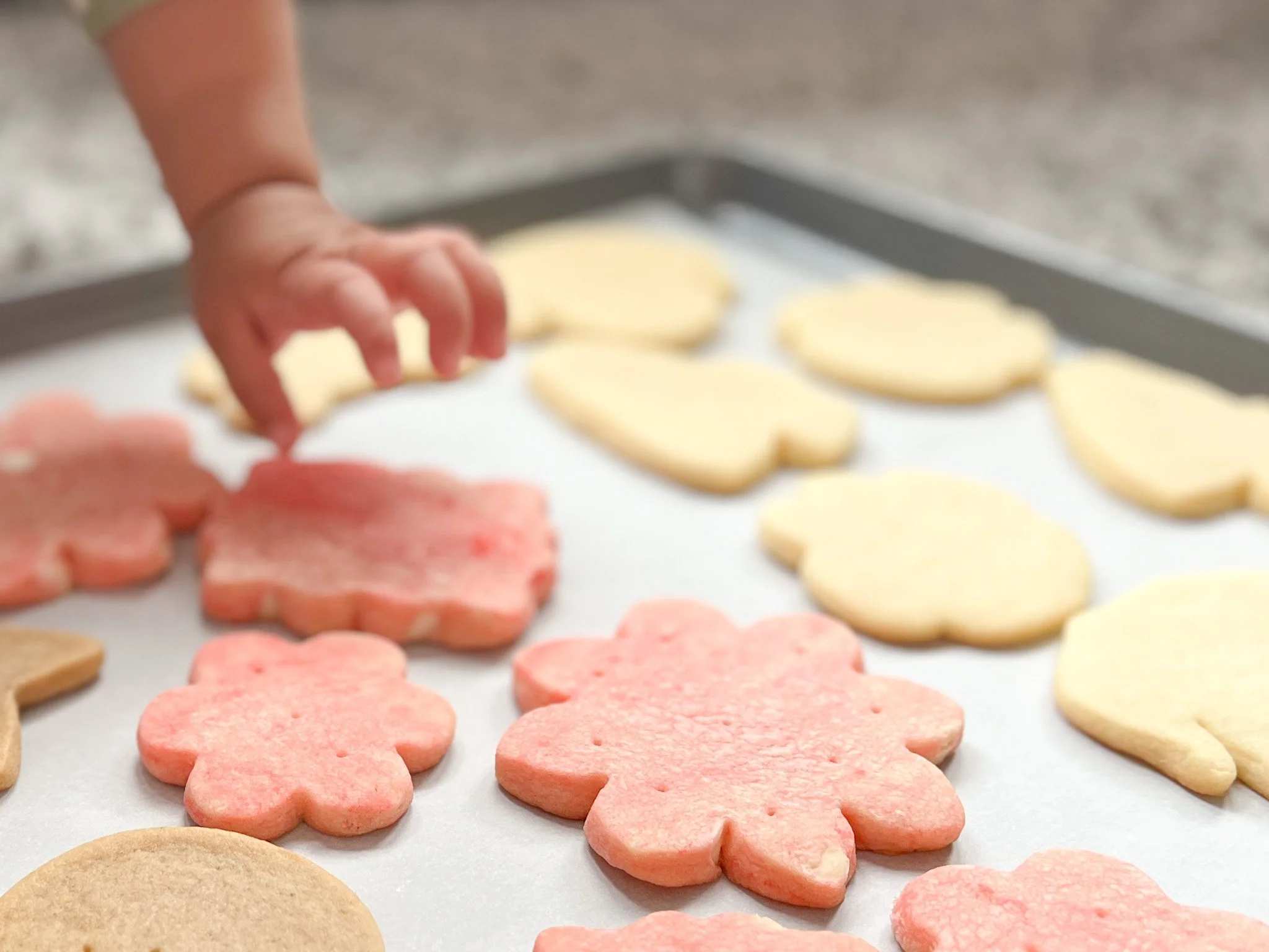 A child's hand reaching for pink and plain shaped cookies on a baking sheet.
