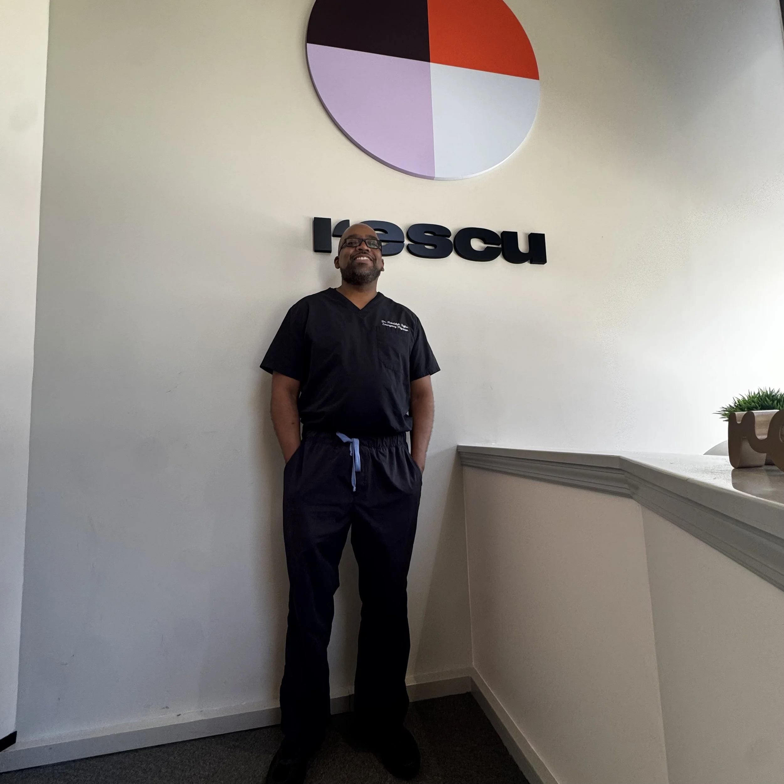 A smiling man with a shaved head and beard wearing a dark scrub top in a medical or clinical setting.