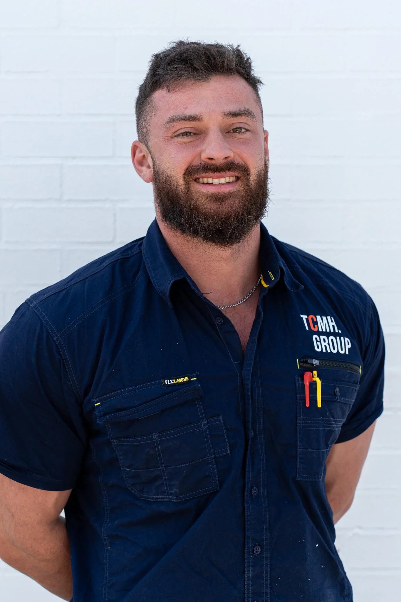 A smiling man with a beard and short hair wearing a navy blue work shirt with patches and pens, standing against a white brick wall.