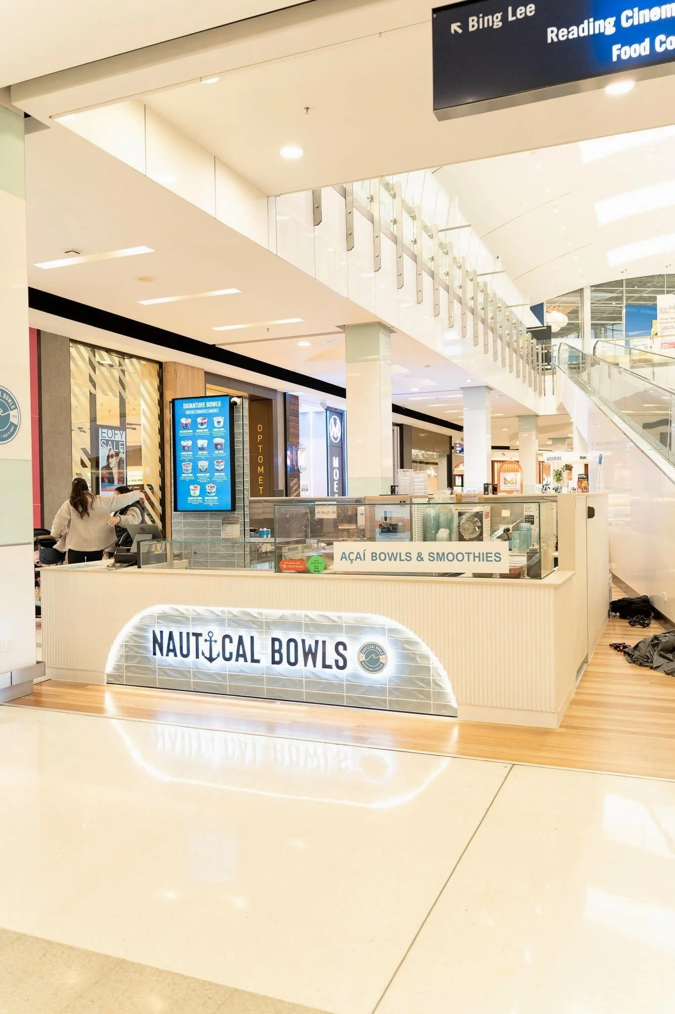 A kiosk selling acai bowls and smoothies named Nautical Bowls inside a shopping mall. The kiosk has a bright sign with an anchor logo and a modern design, with digital menu screens and a counter for orders.
