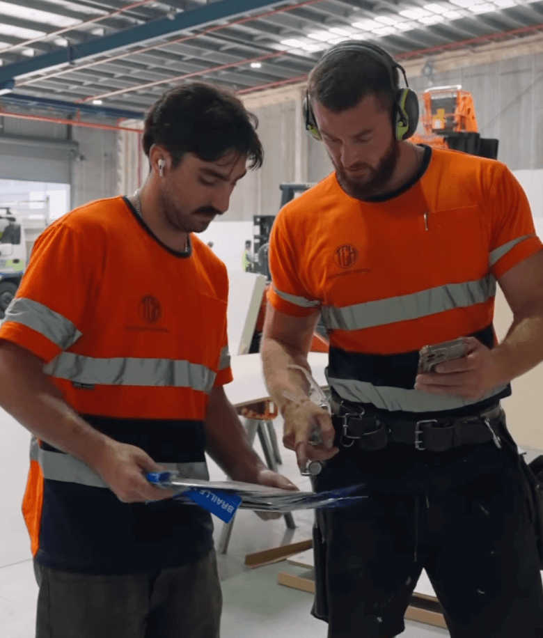 Two construction workers in orange safety shirts with reflective stripes working indoors, looking at documents and a smartphone.