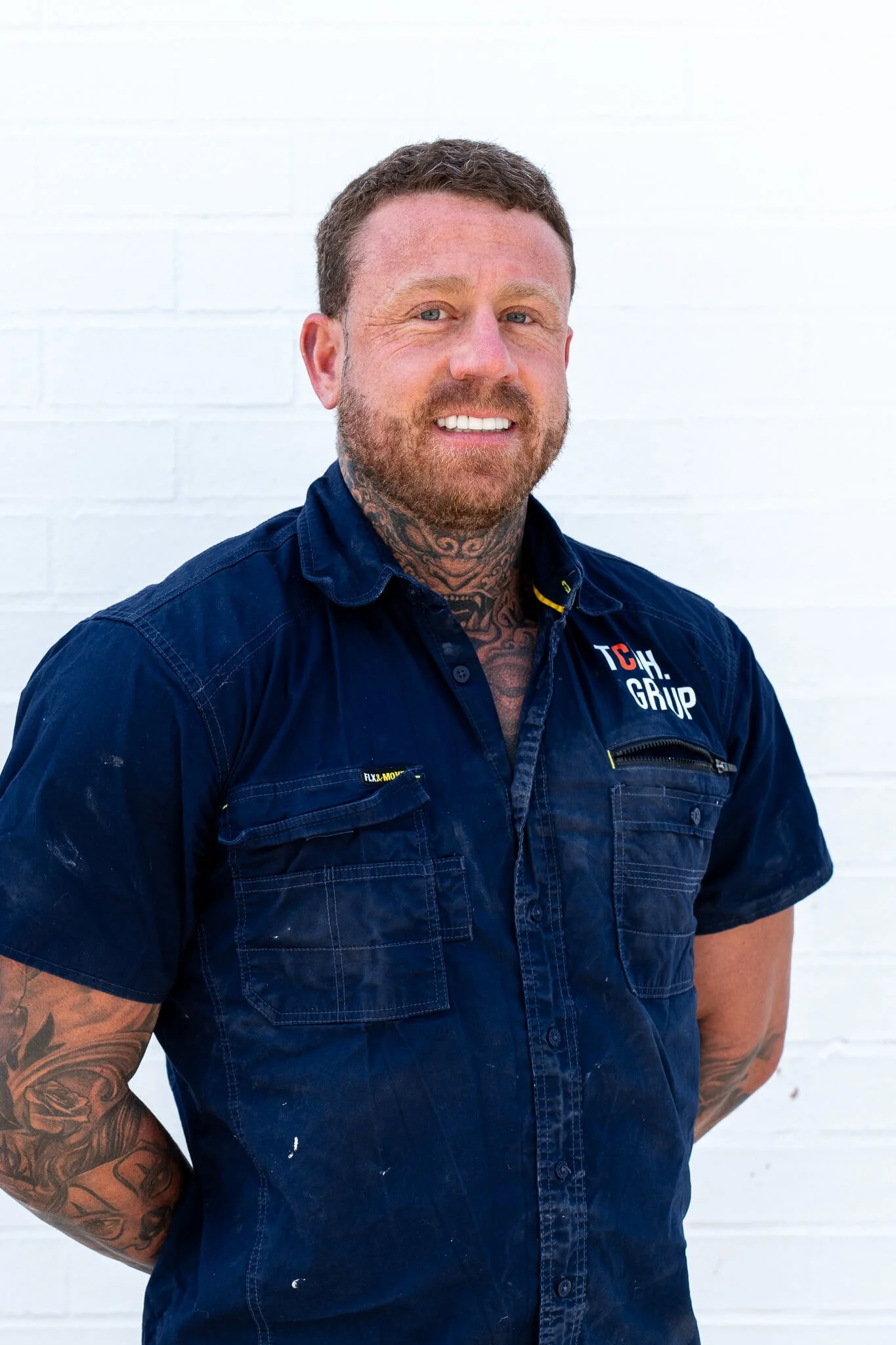 Man with tattoos wearing a blue work shirt with logo, standing against a white brick wall.
