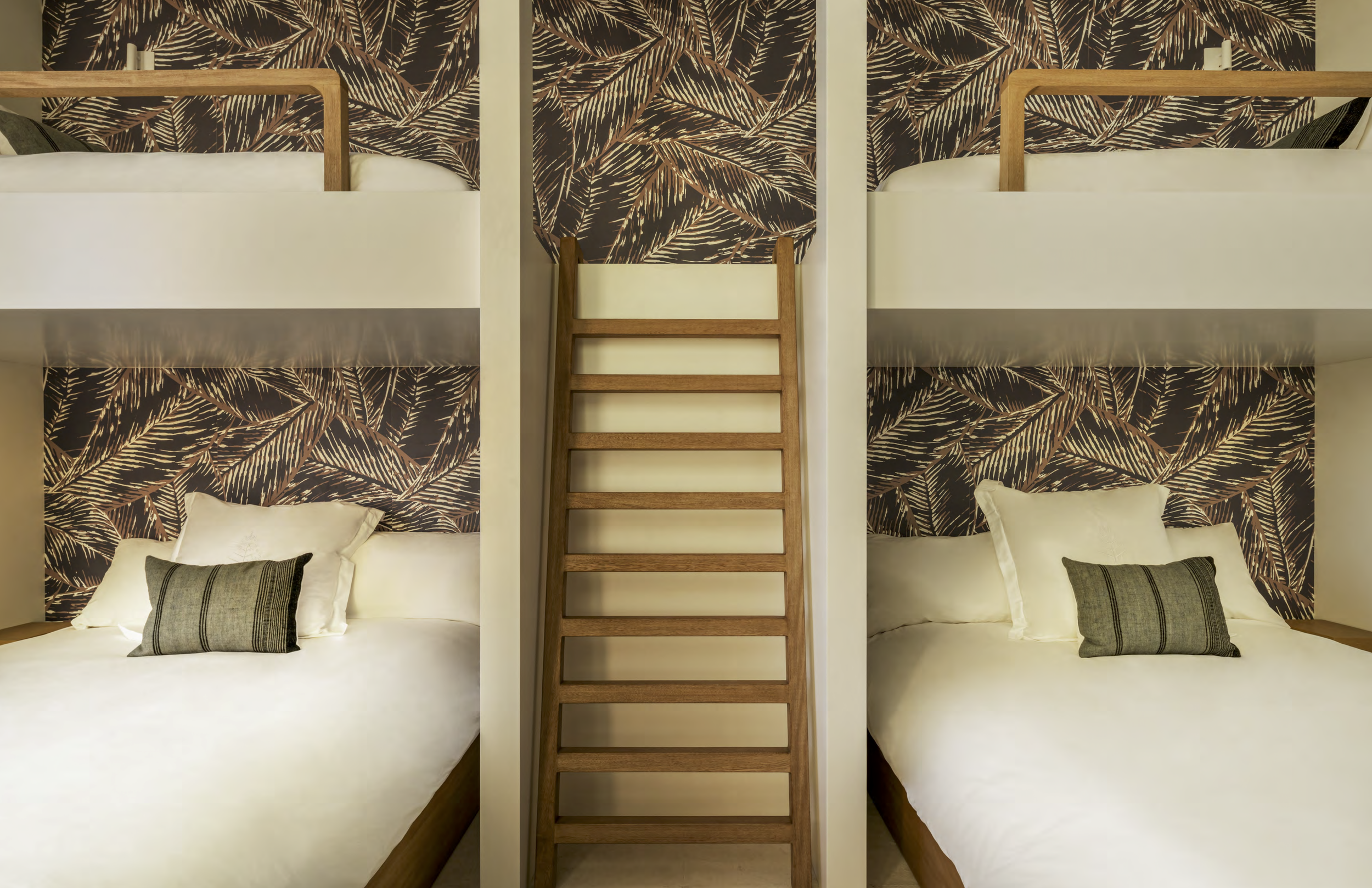 Two adjacent beds in a hotel room with tropical leaf patterned wallpaper behind each bed, a wooden ladder in between, white bedding, and decorative pillows.