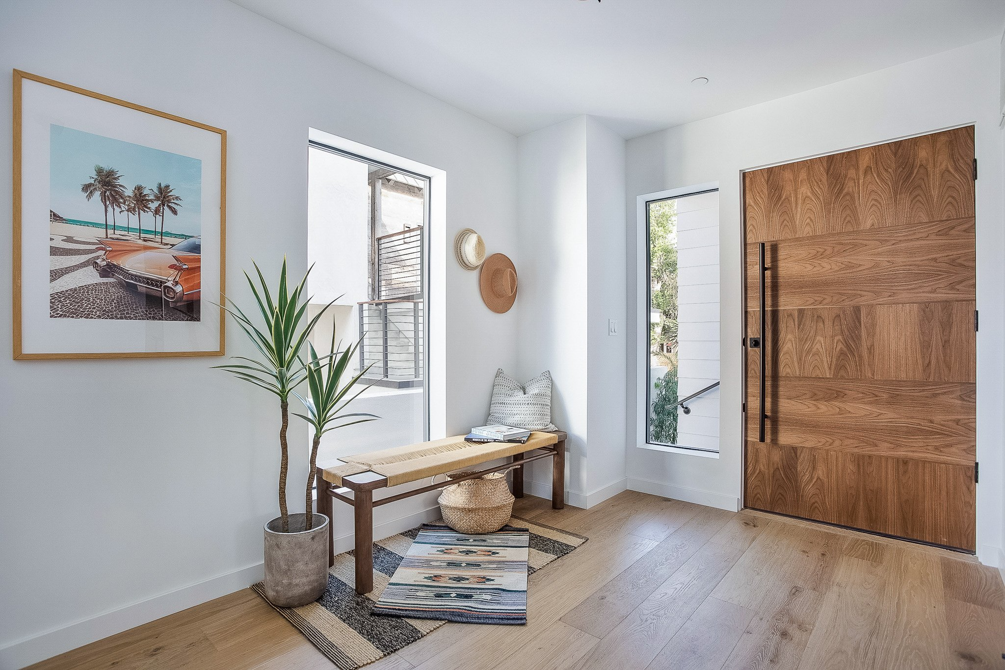 Modern entryway with wooden door, framed beach scene art, potted plant, bench with pillow, hats on wall, and natural light from windows.