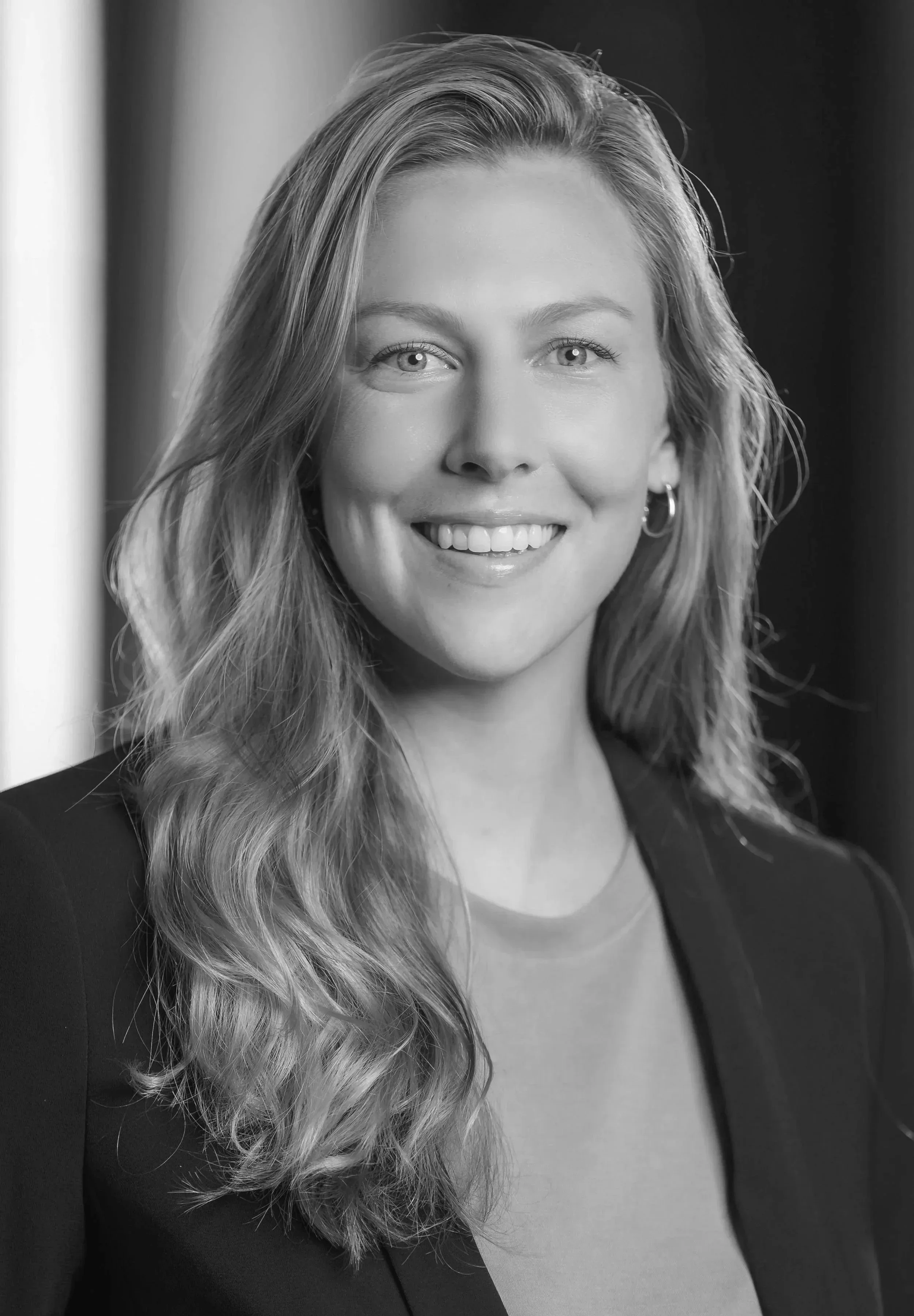 Black and white portrait of a smiling woman with wavy hair, wearing earrings and a blazer.