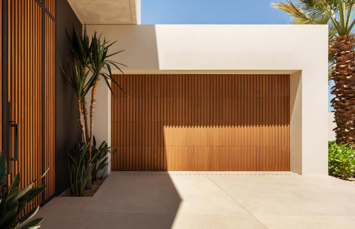 Modern house exterior featuring a wooden garage door, white walls, and desert plants including palm trees and succulents, with a clear blue sky in the background.