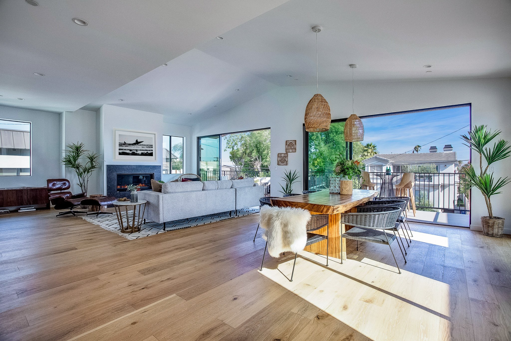 Open-concept living and dining room with hardwood floors, large windows, a gray sofa, a fireplace, a wooden dining table with chairs, and sliding glass doors to a balcony with outdoor seating and greenery.