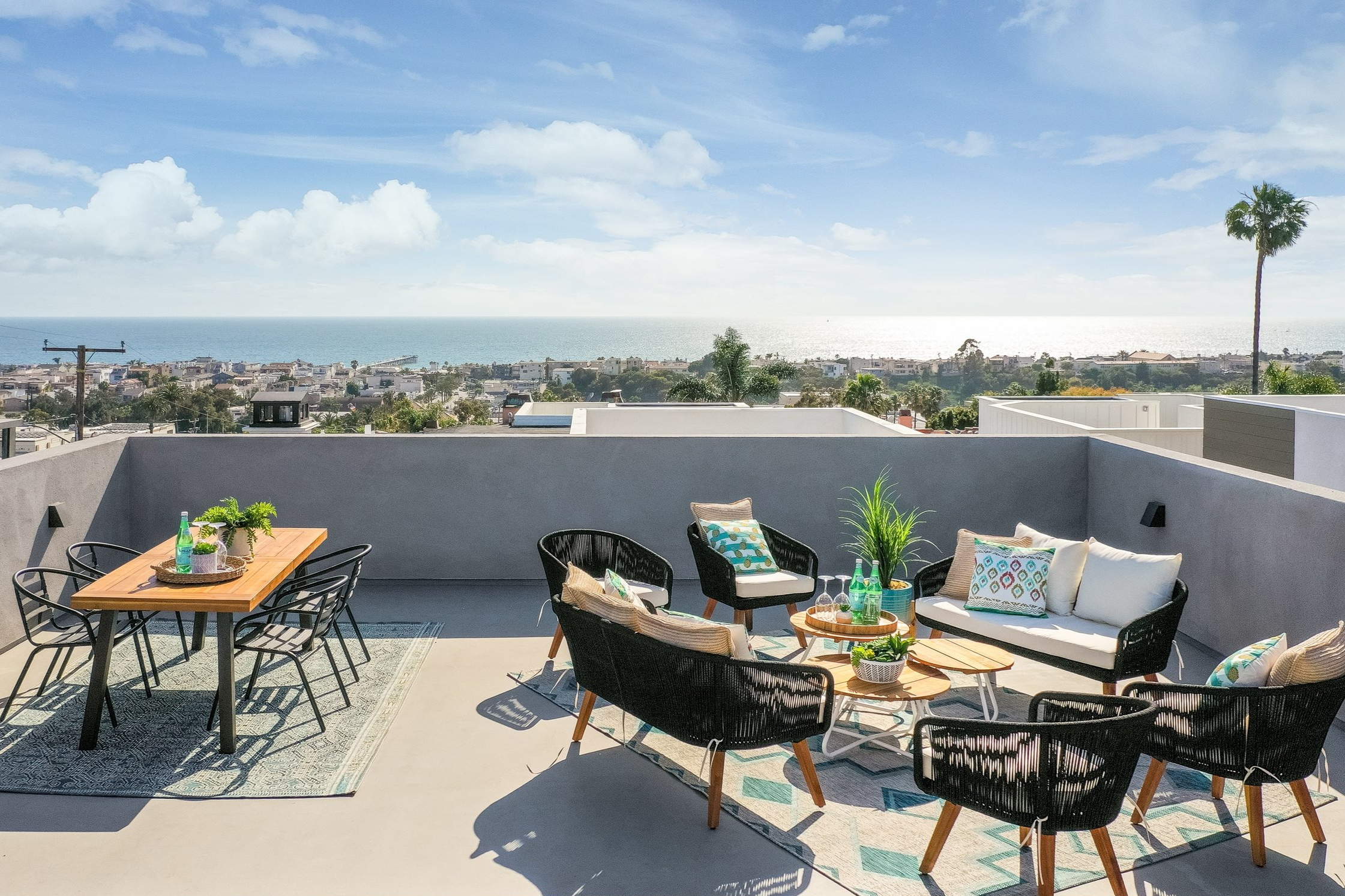 Outdoor rooftop patio with black wicker chairs, white sofa with decorative pillows, wooden tables, and potted plants, overlooking a cityscape with the ocean in the distance.