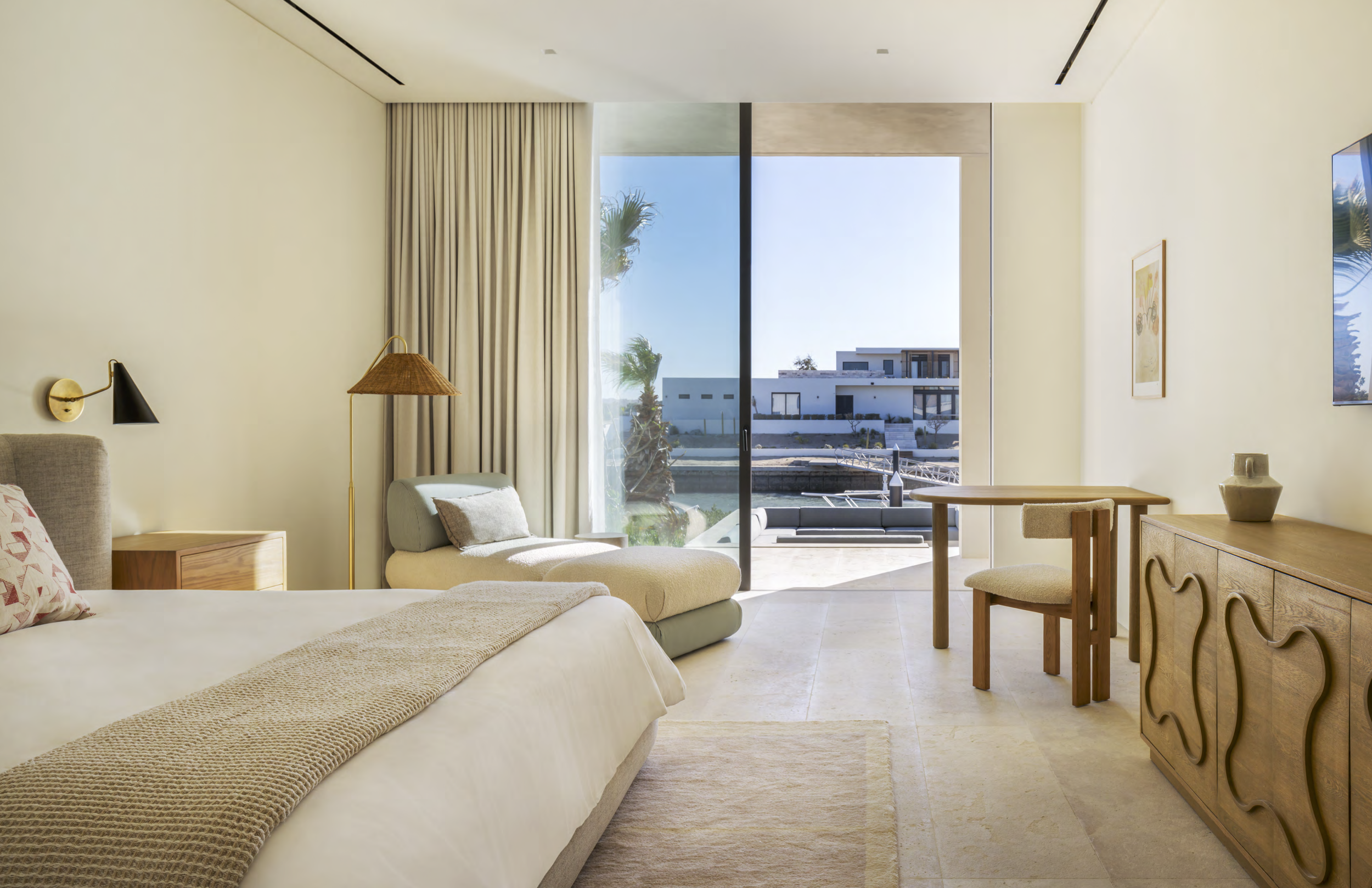 Bright hotel room with a large bed, beige and gray furniture, a sliding glass door leading to a balcony with a view of modern buildings, palm trees, and water, and a wooden sideboard.