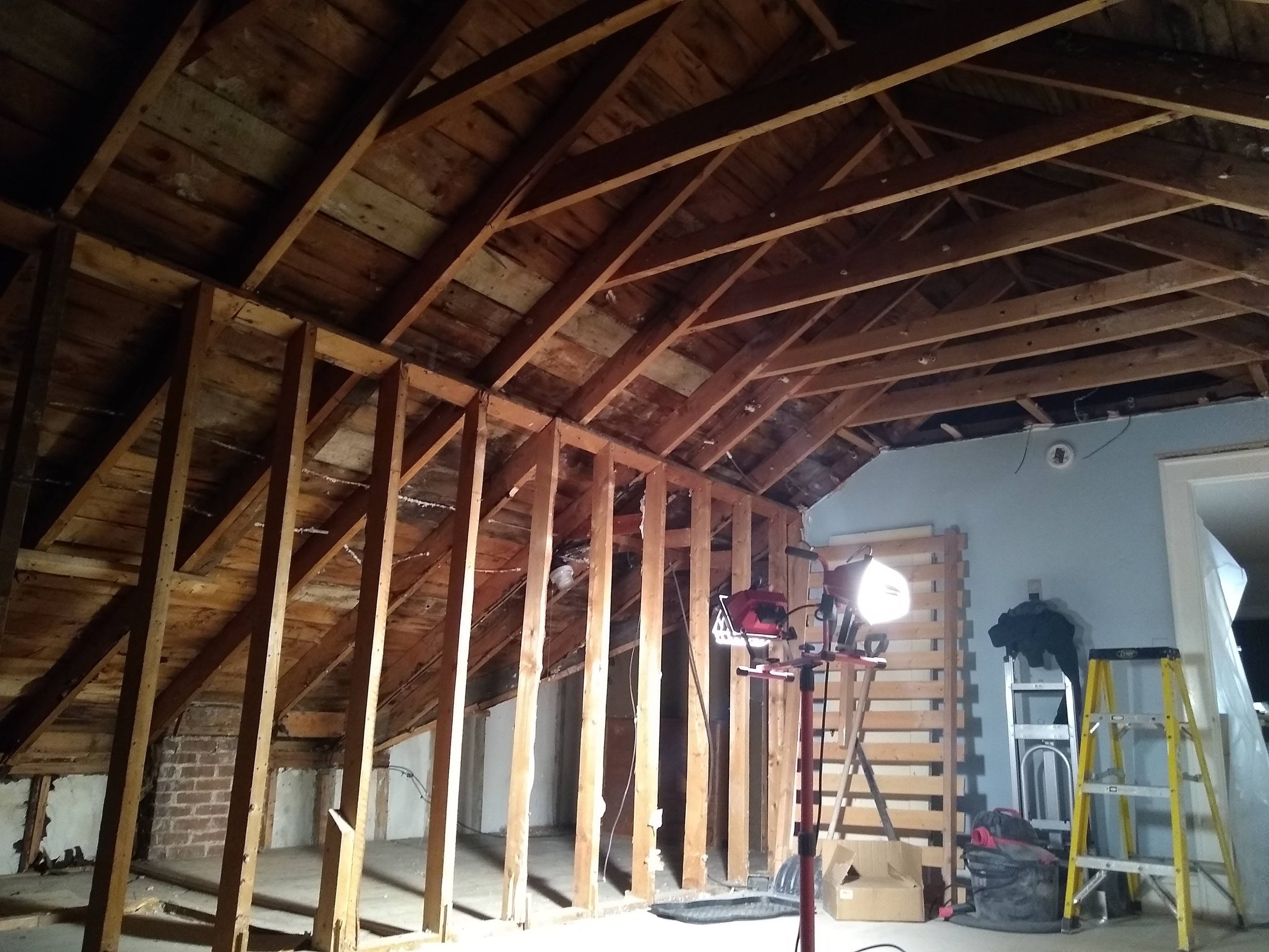 Interior of a house under renovation with exposed wooden framing, a ladder, work light, and construction tools.