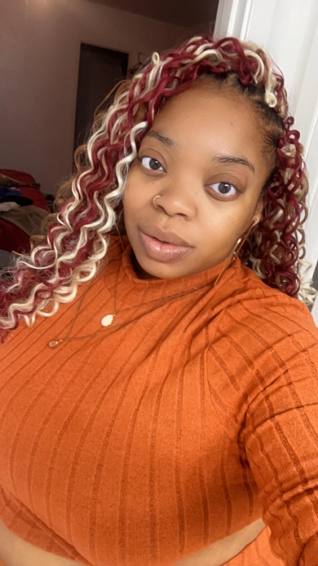 Person with curly red and blonde hair, wearing an orange sweater and necklace in a room.