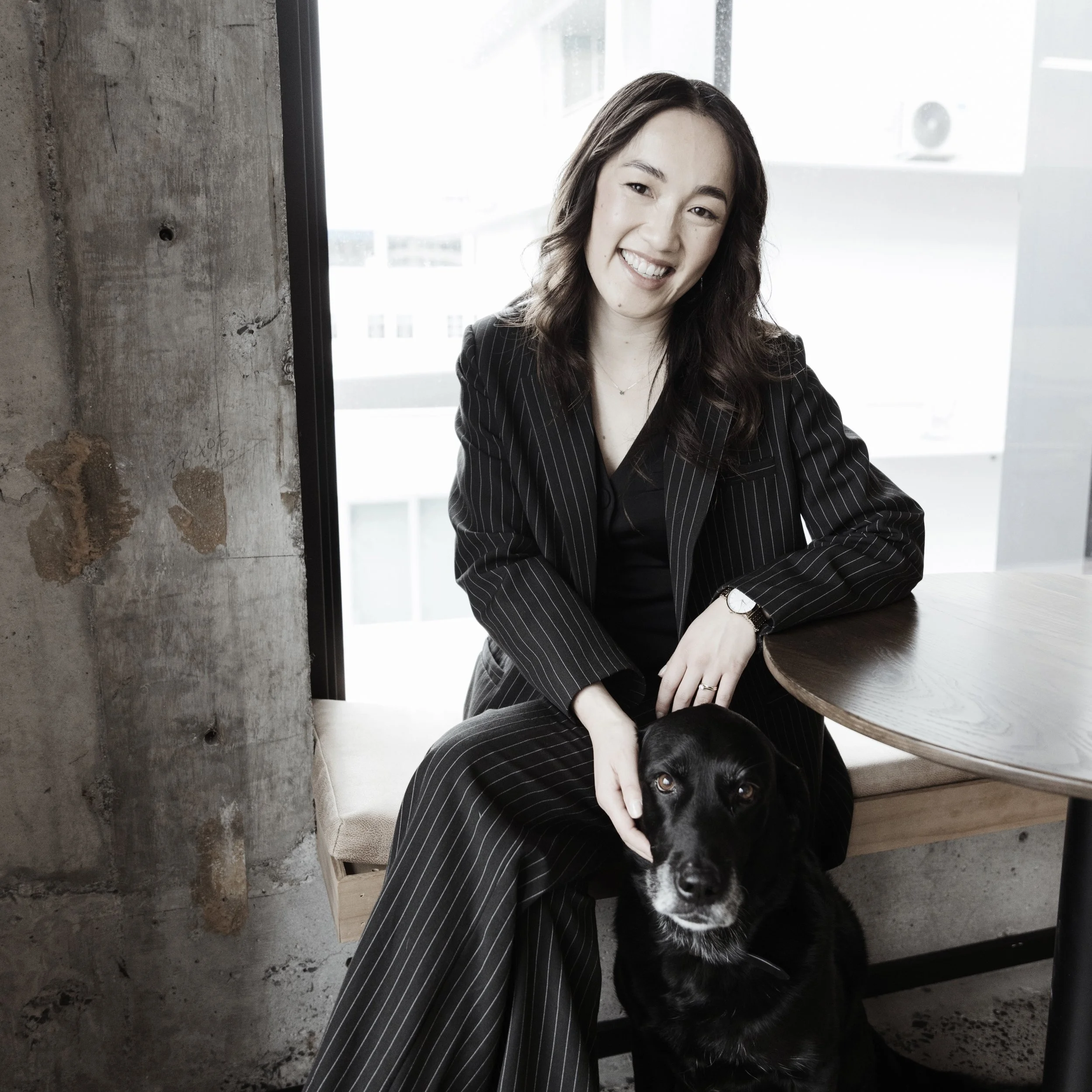 Janey Haringa - a woman with wavy brown hair wearing a black pinstripe suit sitting on a bench with a black dog, smiling at the camera indoors by a window.