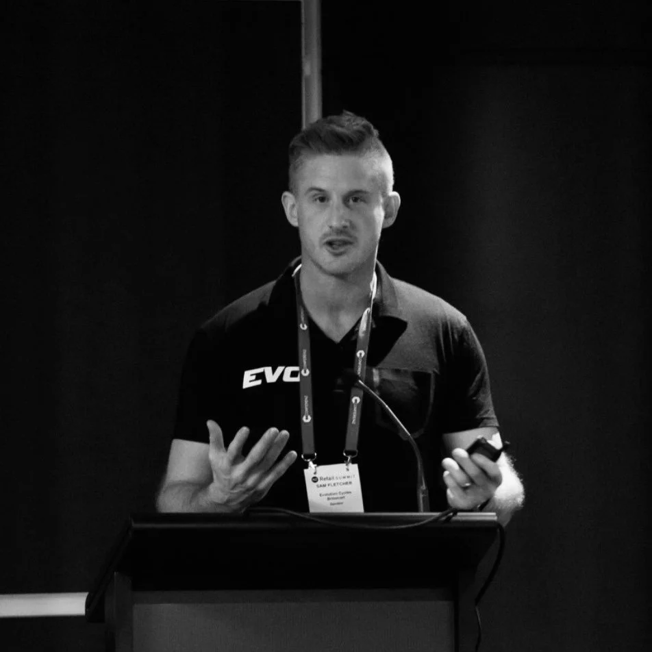 Sam Fletcher giving a presentation at a conference. He is wearing a black shirt with his company logo 'EVO' and has a conference badge around his neck. He is gesturing with one hand and holding a device, possibly a remote or phone, in the other.