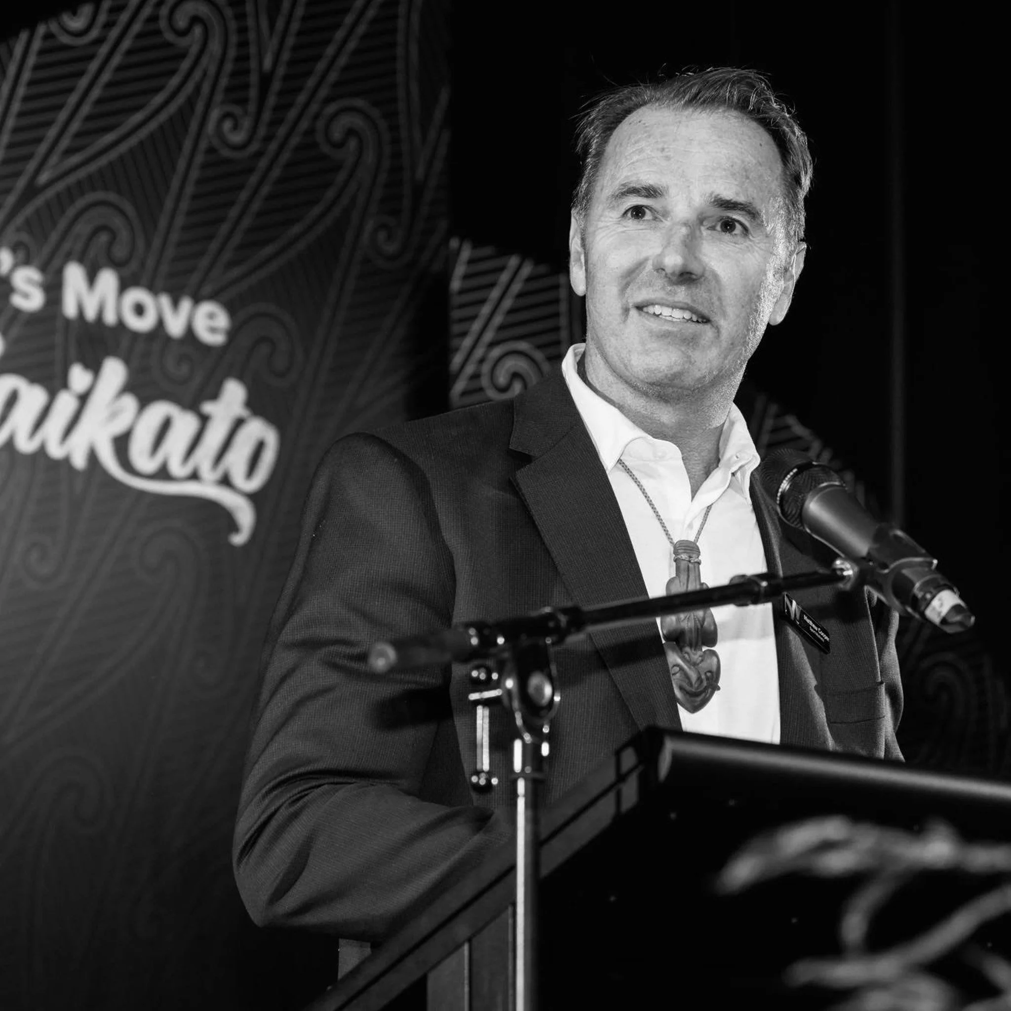 Matthew Cooper (President of New Zealand Rugby and Chief Executive of Sport Waikato) in a suit and white shirt speaking at a podium with a microphone, with a dark background and a sign that reads 'Let's Move Waikato.'