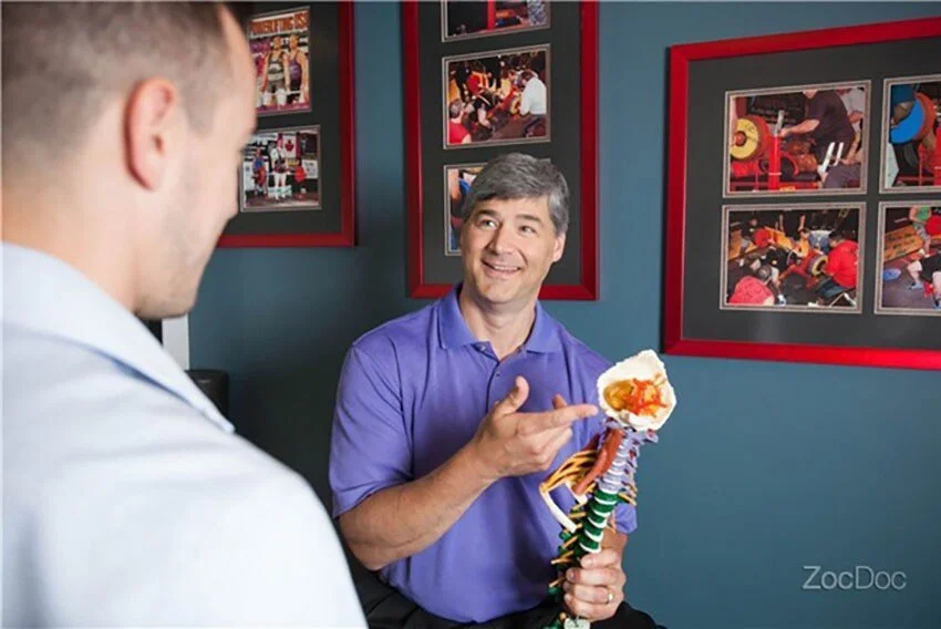 Dr. Mark Webber, chiropractor, in a purple shirt holds a model of a spine, explaining to another person in the Kirkland, WA clinic with framed pictures on the wall.