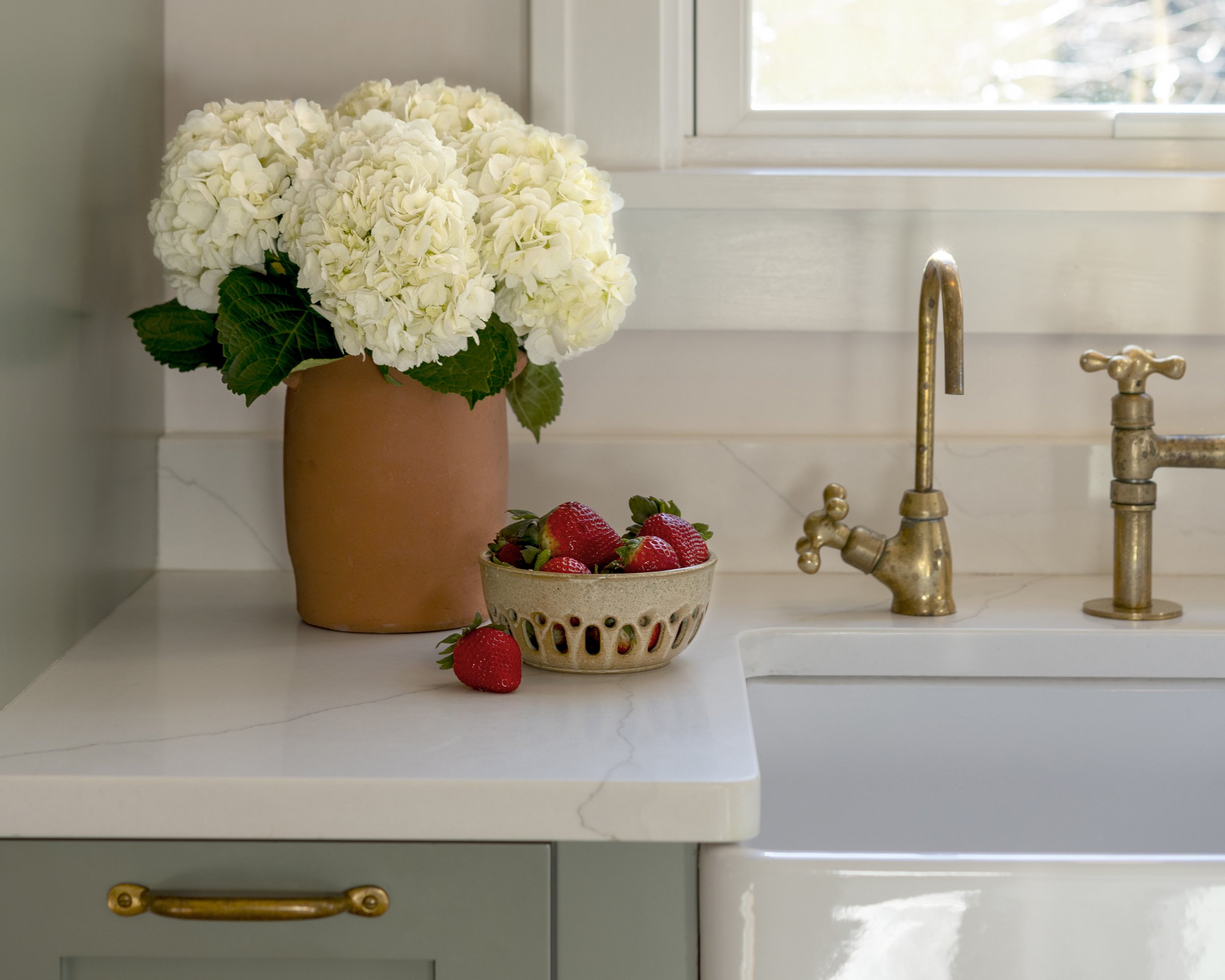 White hydrangea flowers in a terracotta pot, a bowl of strawberries on a white kitchen counter near a brass faucet, with a window in the background.