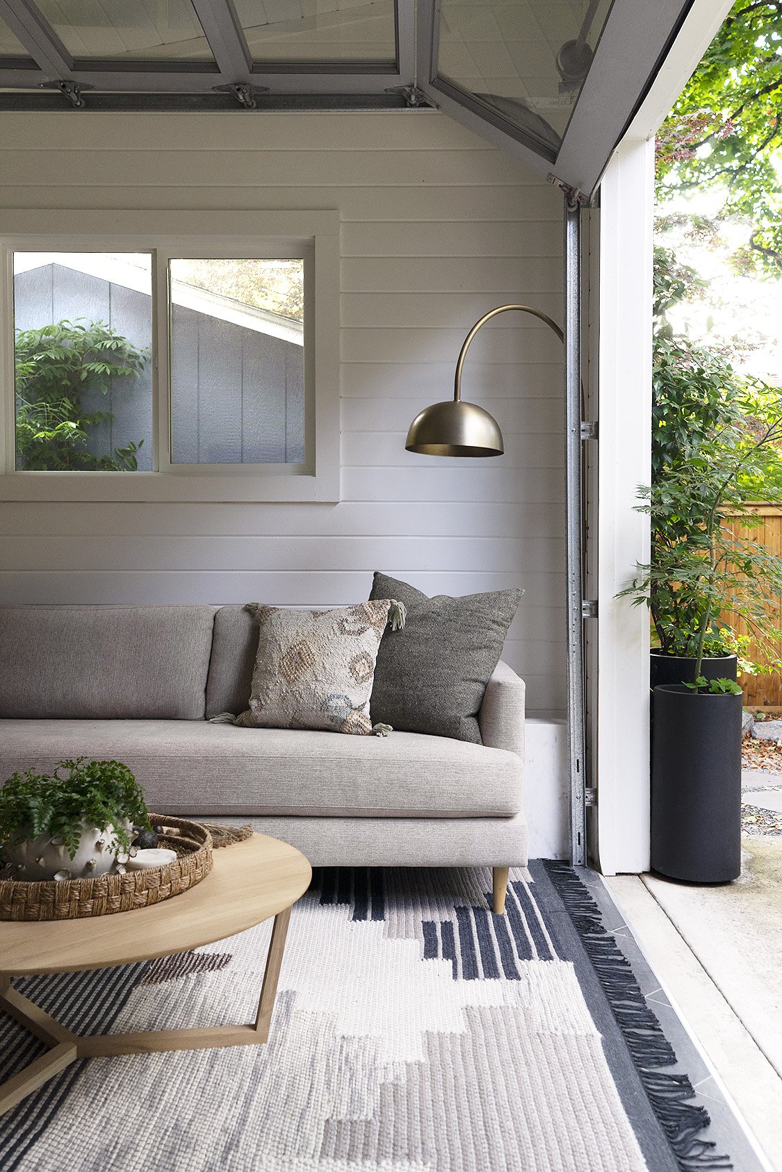A cozy living room with a beige sofa, decorative pillows, a wooden coffee table with a potted plant, a wall-mounted brass reading lamp, a window, and an open garage door leading outside with greenery and a wooden fence.