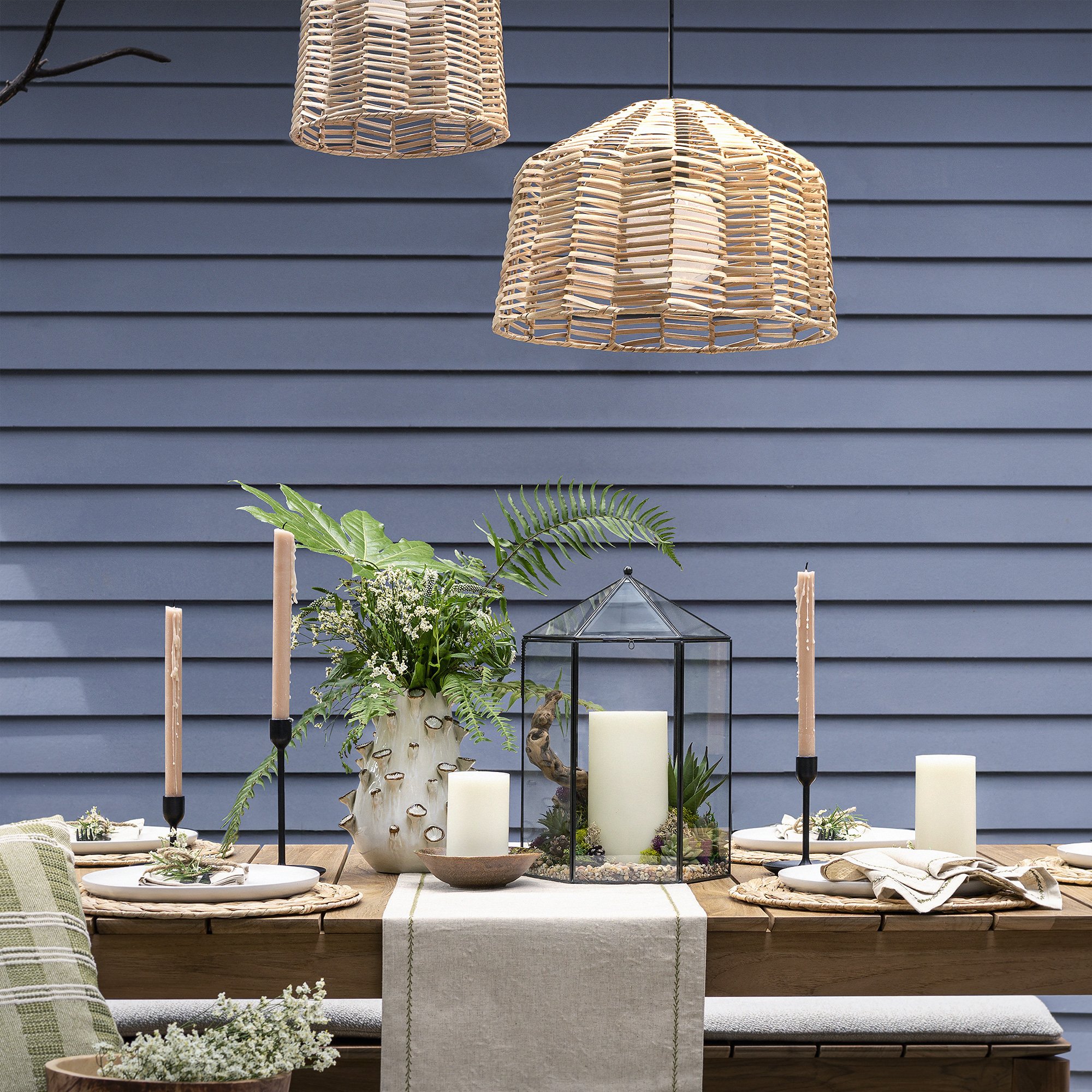A dining table decorated with candles, green plants, and a glass terrarium, set against a dark blue horizontal siding wall. Two woven rattan pendant lights hang above the table.