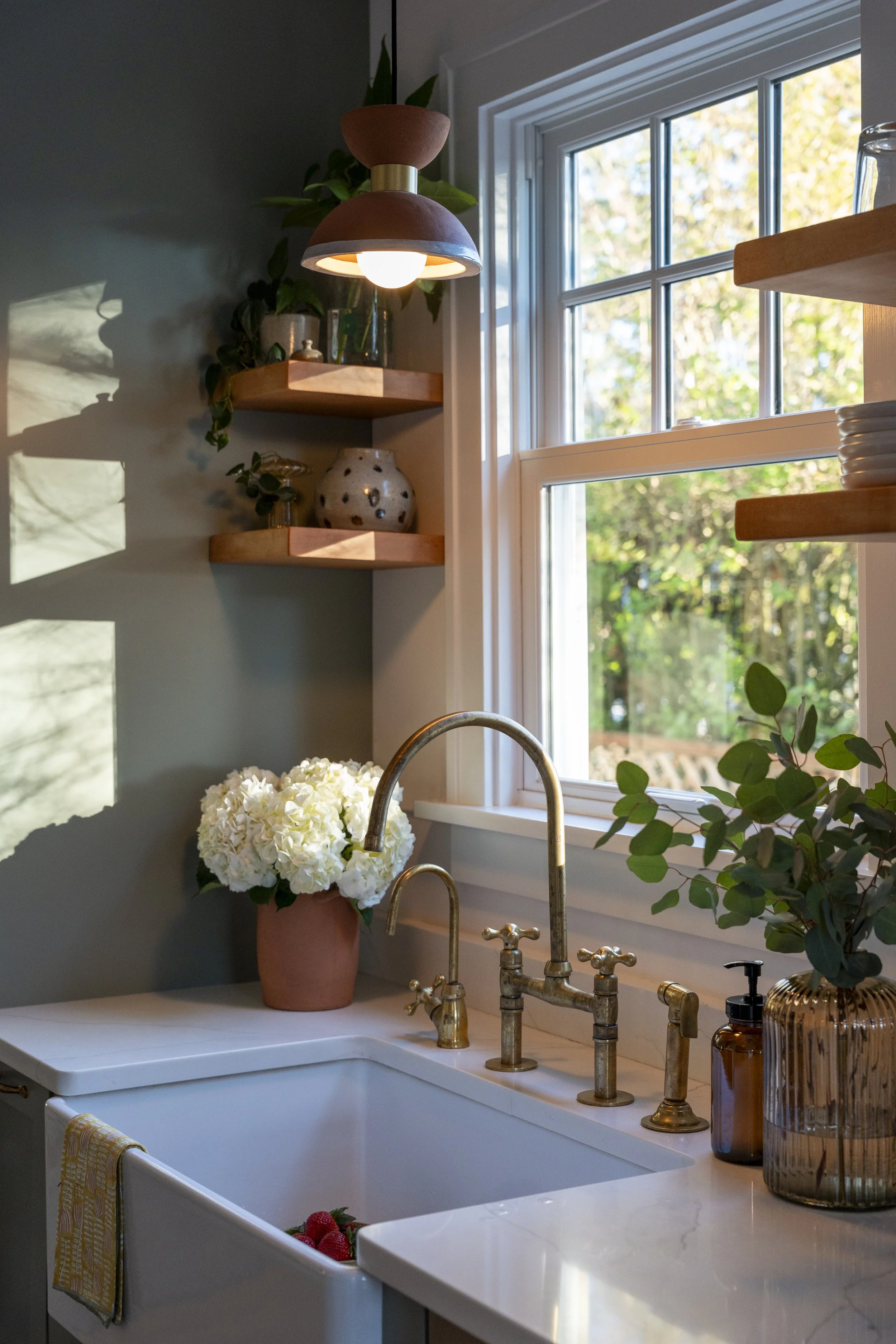 Kitchen window with a view of trees and sunlight, a white farmhouse sink with a brass faucet, a potted hydrangea flower, and decorative items on wooden shelves including a pottery vase, a glass jar with a plant, and amber-colored containers.