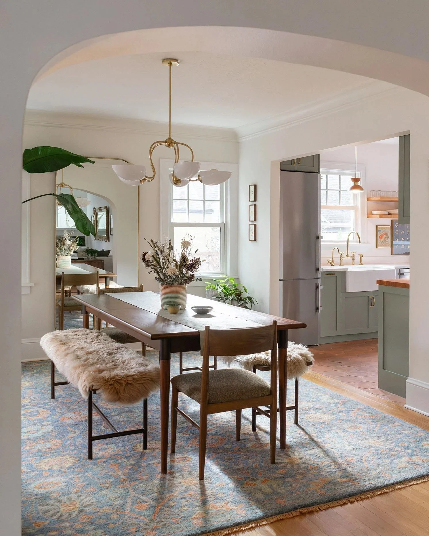 Dining room with wooden table, two chairs, a fluffy bench, a vase with dried flowers, a chandelier, and window with sunlight, opening to the kitchen with pastel green cabinets and a refrigerator.