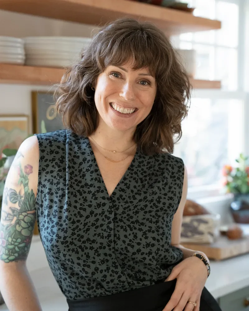 A woman with curly brown hair smiling, wearing a sleeveless black dress with a floral pattern, standing in a bright kitchen with wooden shelves, plants, and framed art in the background.