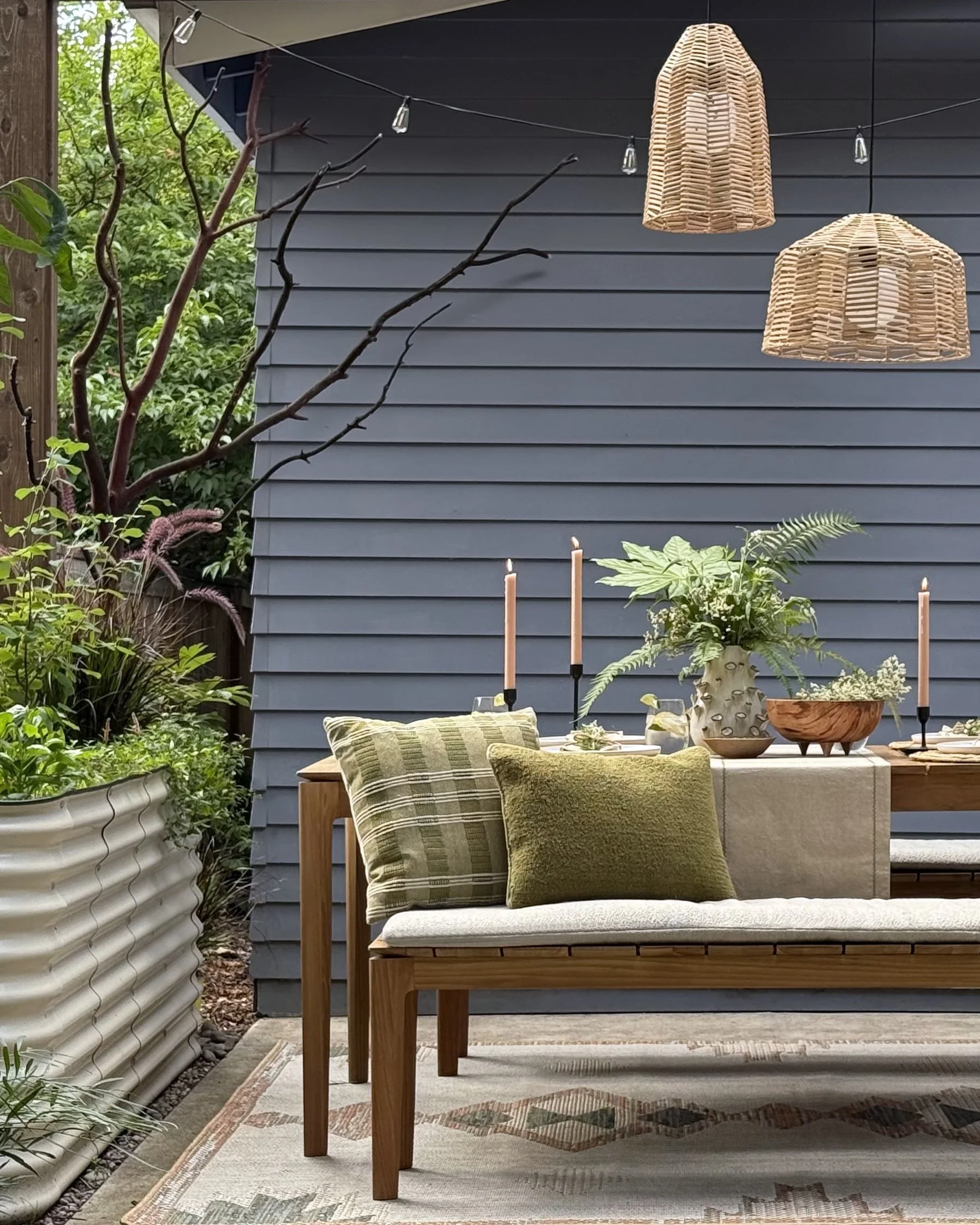 Outdoor dining area with wooden table, green and patterned pillows on seat, large potted plant, two wicker hanging lamps, and greenery in the background.